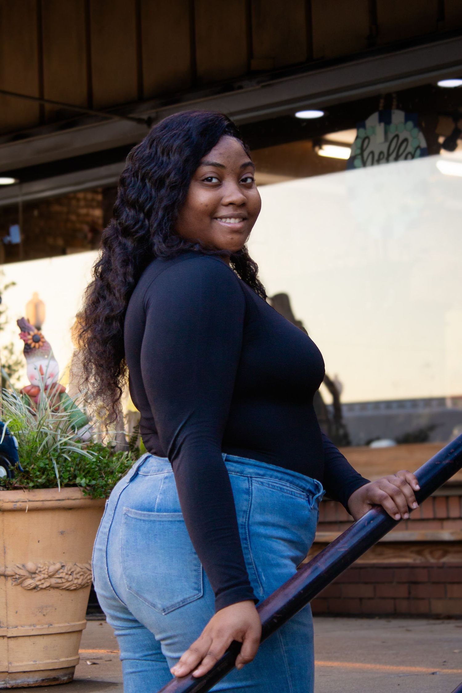 Smiling young Black woman in black top and jeans poses by railing outside a shop.