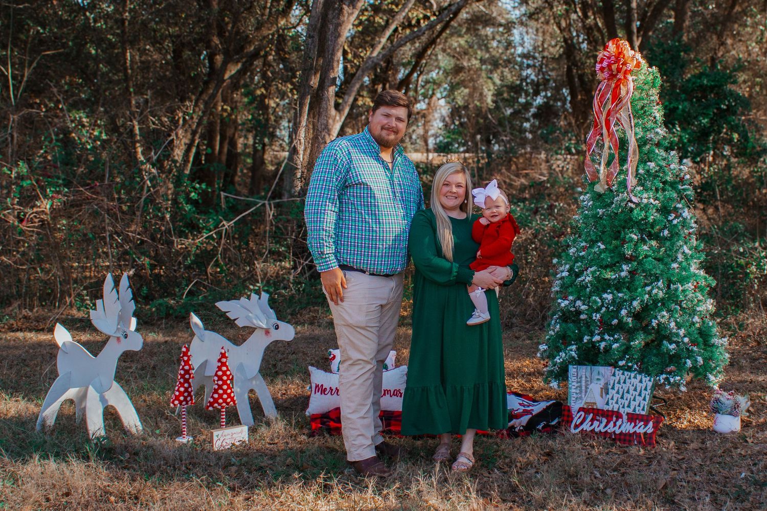 Family of three posing by a Christmas tree with wooden reindeer decorations in an outdoor holiday setting.