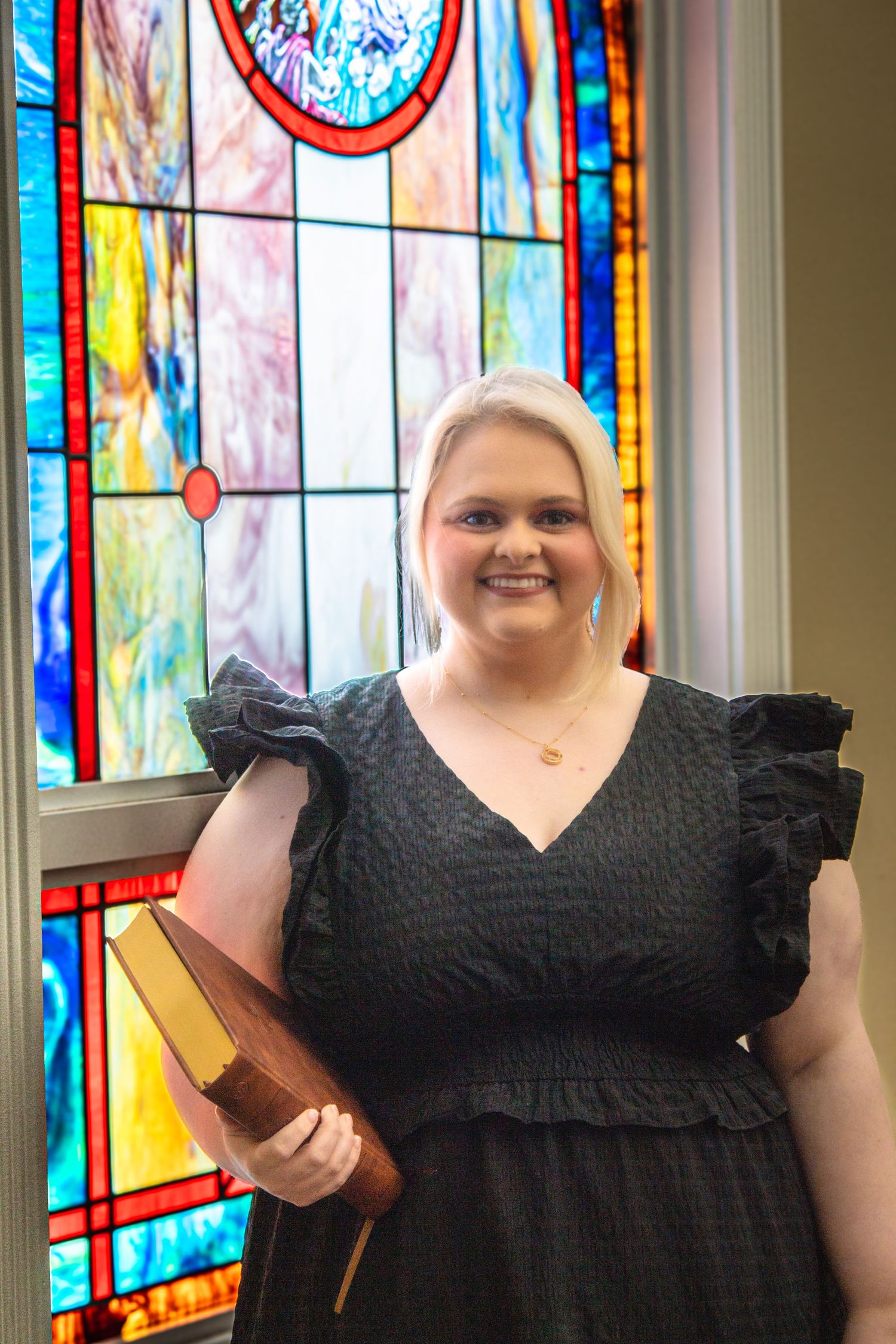 Blonde woman in black ruffled dress holding a Bible while standing before a colorful stained glass window.