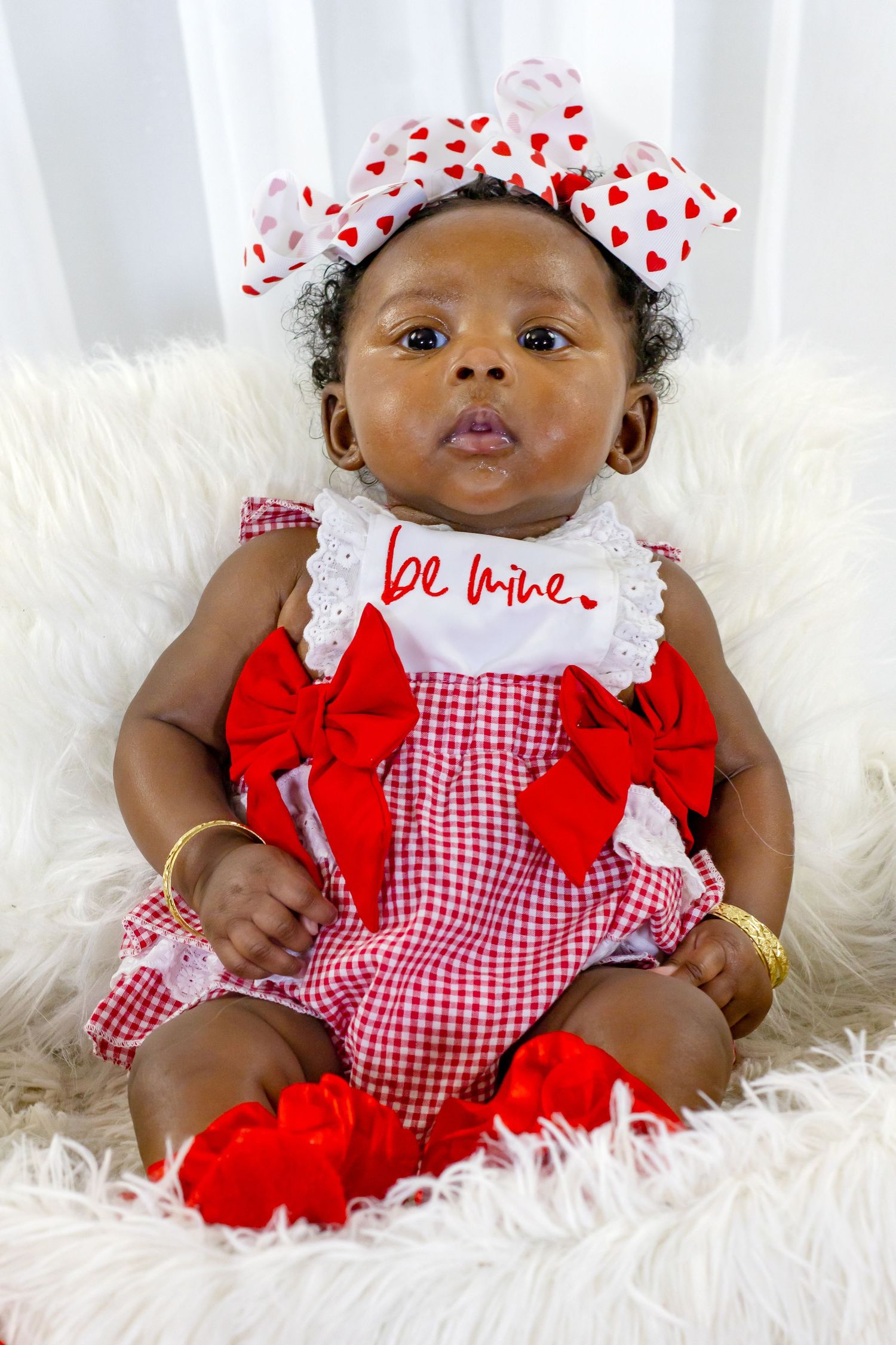 Adorable Black baby in red gingham Be Mine outfit with bow headband on white fur.
