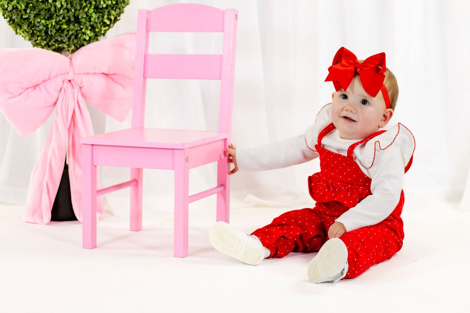 Baby girl in red polka dot outfit and bow headband sits beside a pink chair with Valentine decorations.