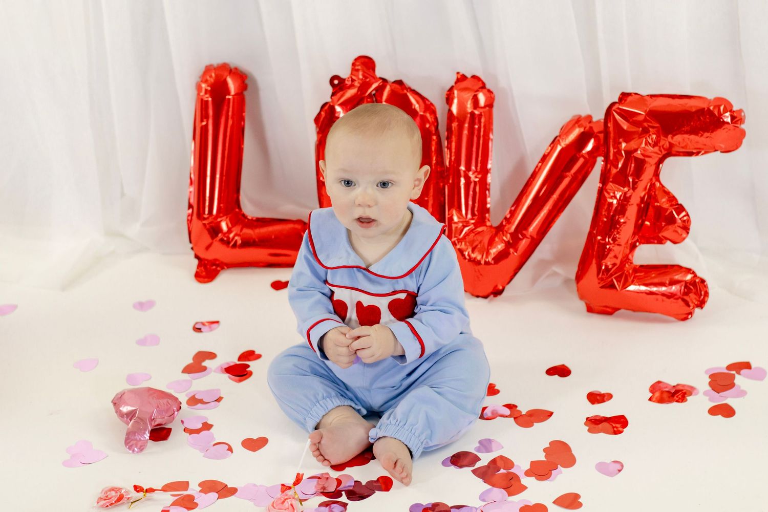 Baby in blue heart romper sits before red LOVE balloon letters surrounded by heart confetti for Valentine's Day.