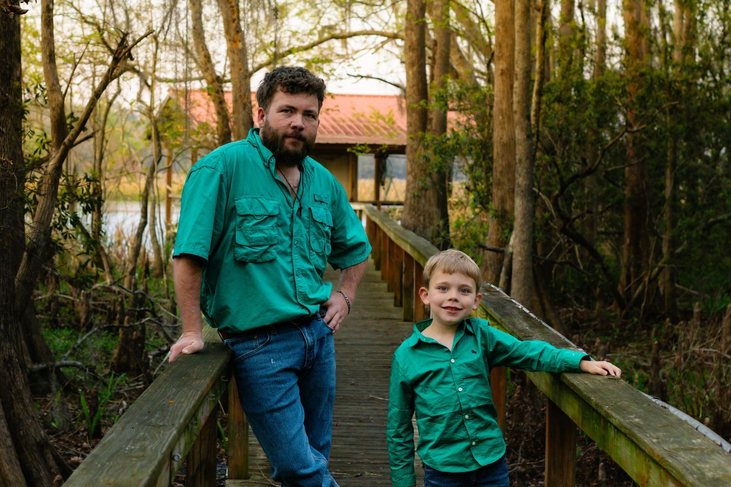 Father and son in matching green shirts pose together on a wooden bridge in a wooded area.