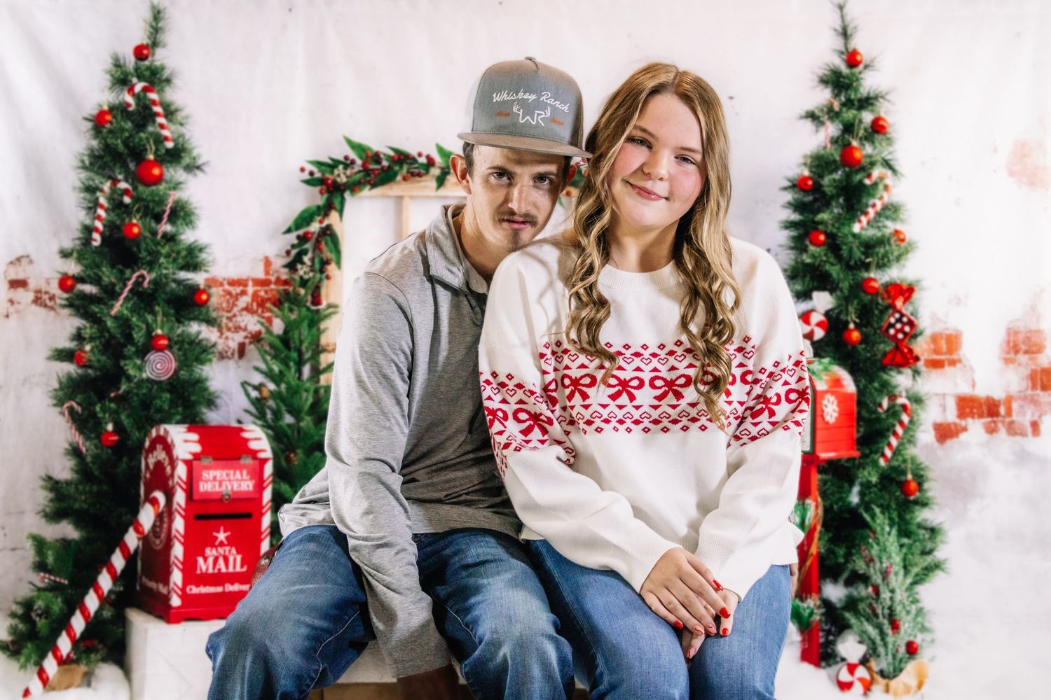 A couple in matching holiday sweaters poses together in front of decorated Christmas trees in a studio portrait.