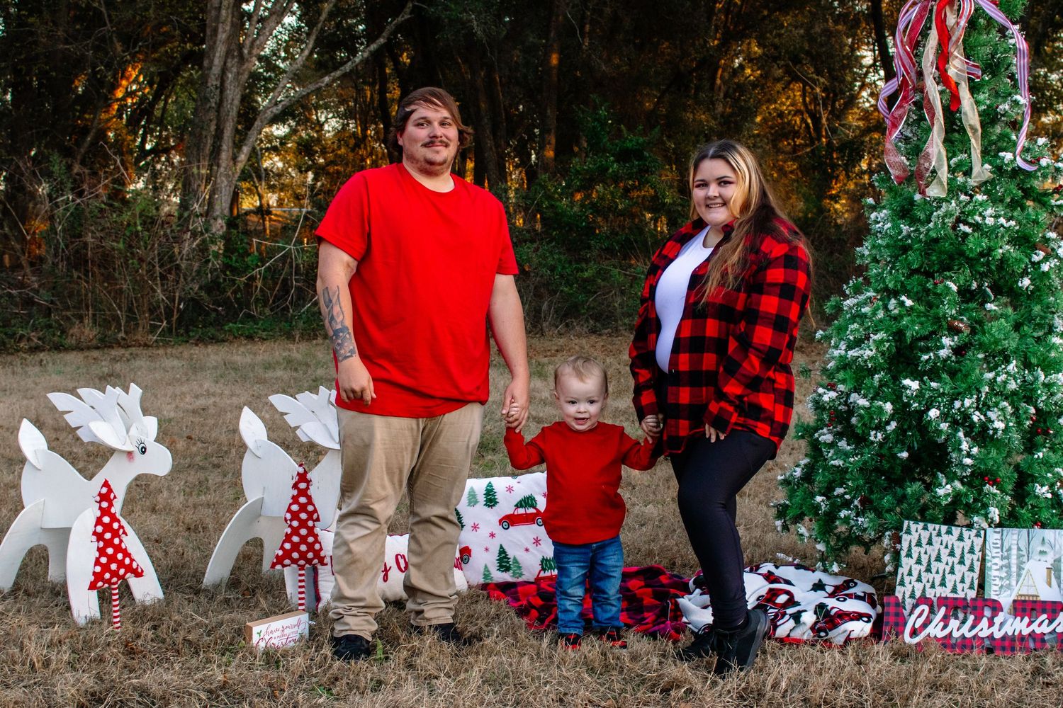 Family of three in red outfits posing outdoors beside a white Christmas tree with wrapped gifts and reindeer decorations.