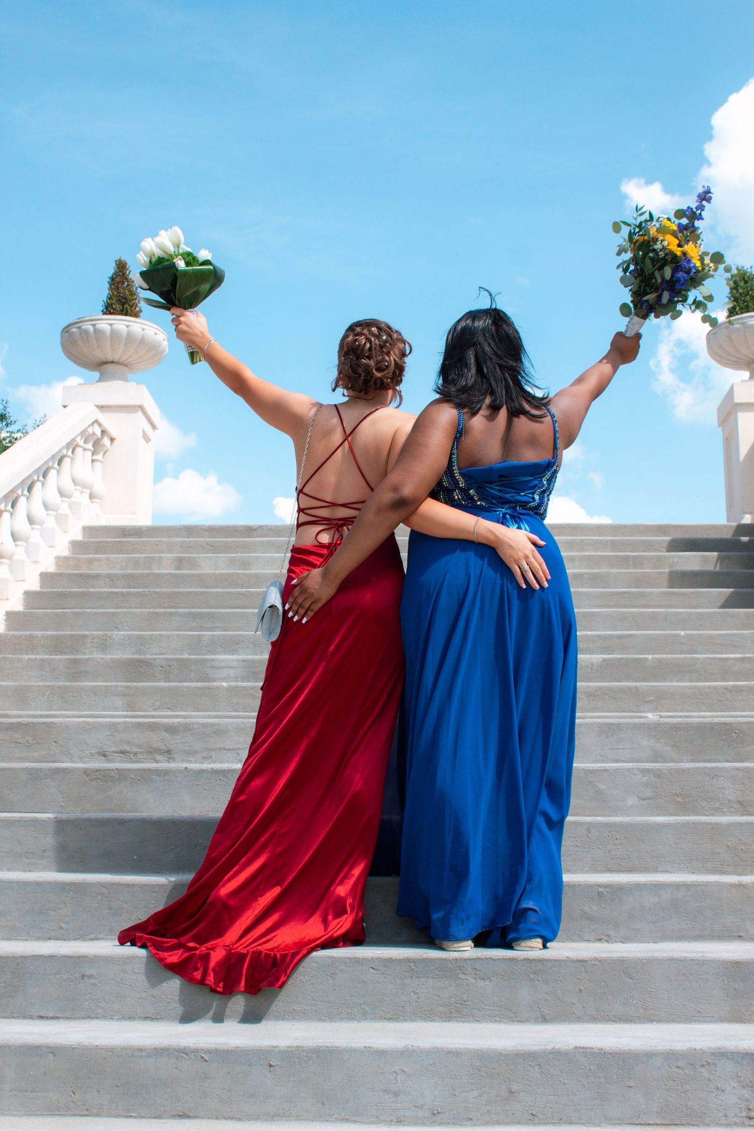 Two prom girls in red and blue gowns raise their bouquets triumphantly while ascending white outdoor stairs.