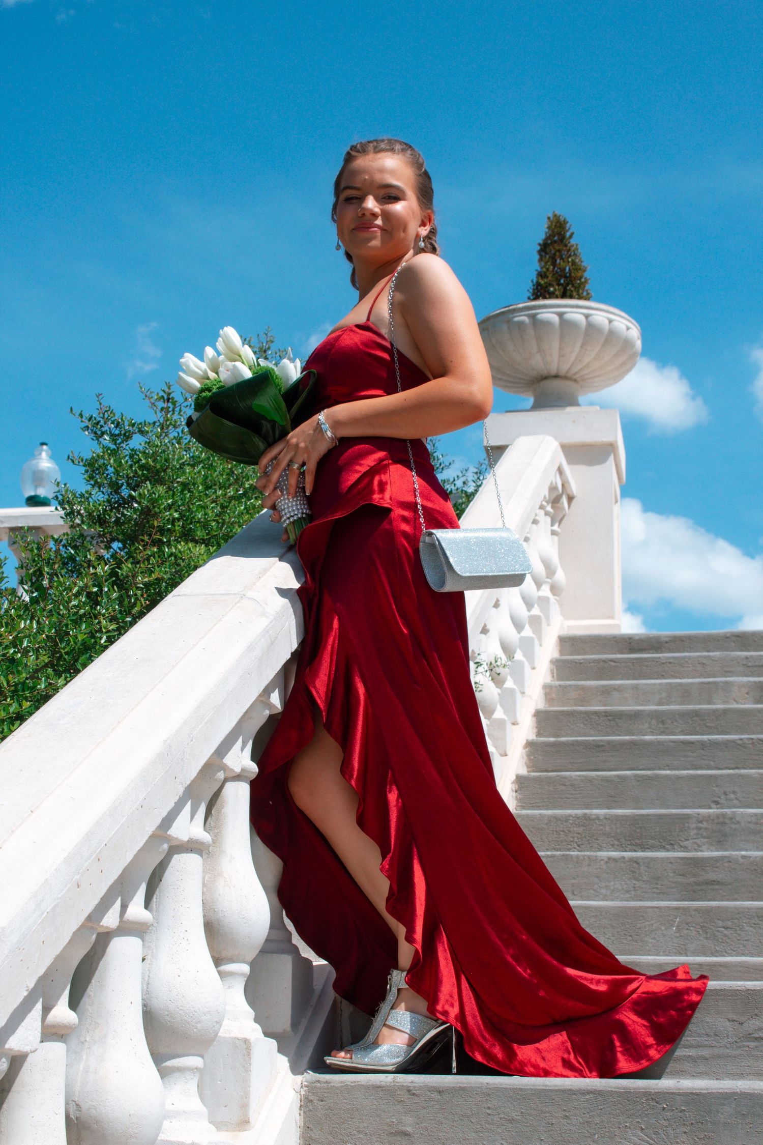 A young woman in a red formal gown holds white tulips while posing on white stairs.