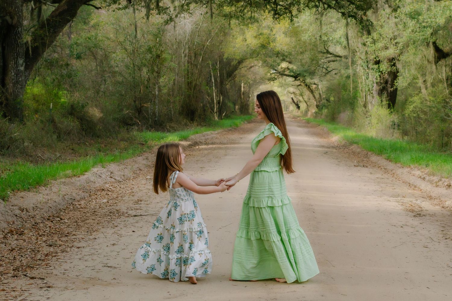 Two girls in green dresses hold hands and dance on a wooded dirt road.