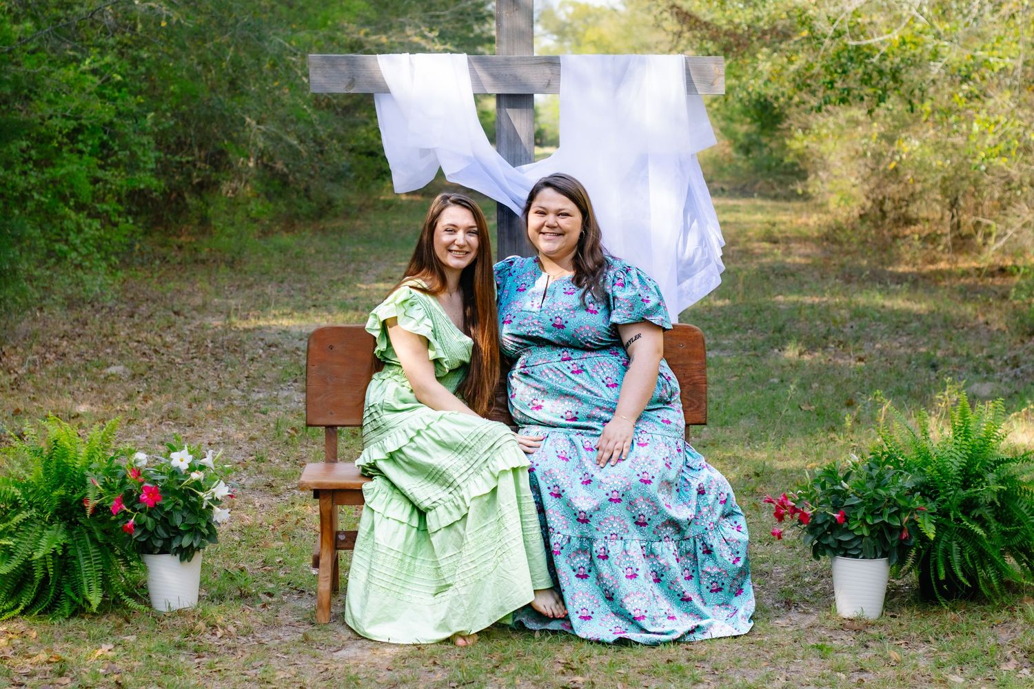 Two young women in floral dresses sitting on a wooden bench with potted flowers and a cross backdrop.
