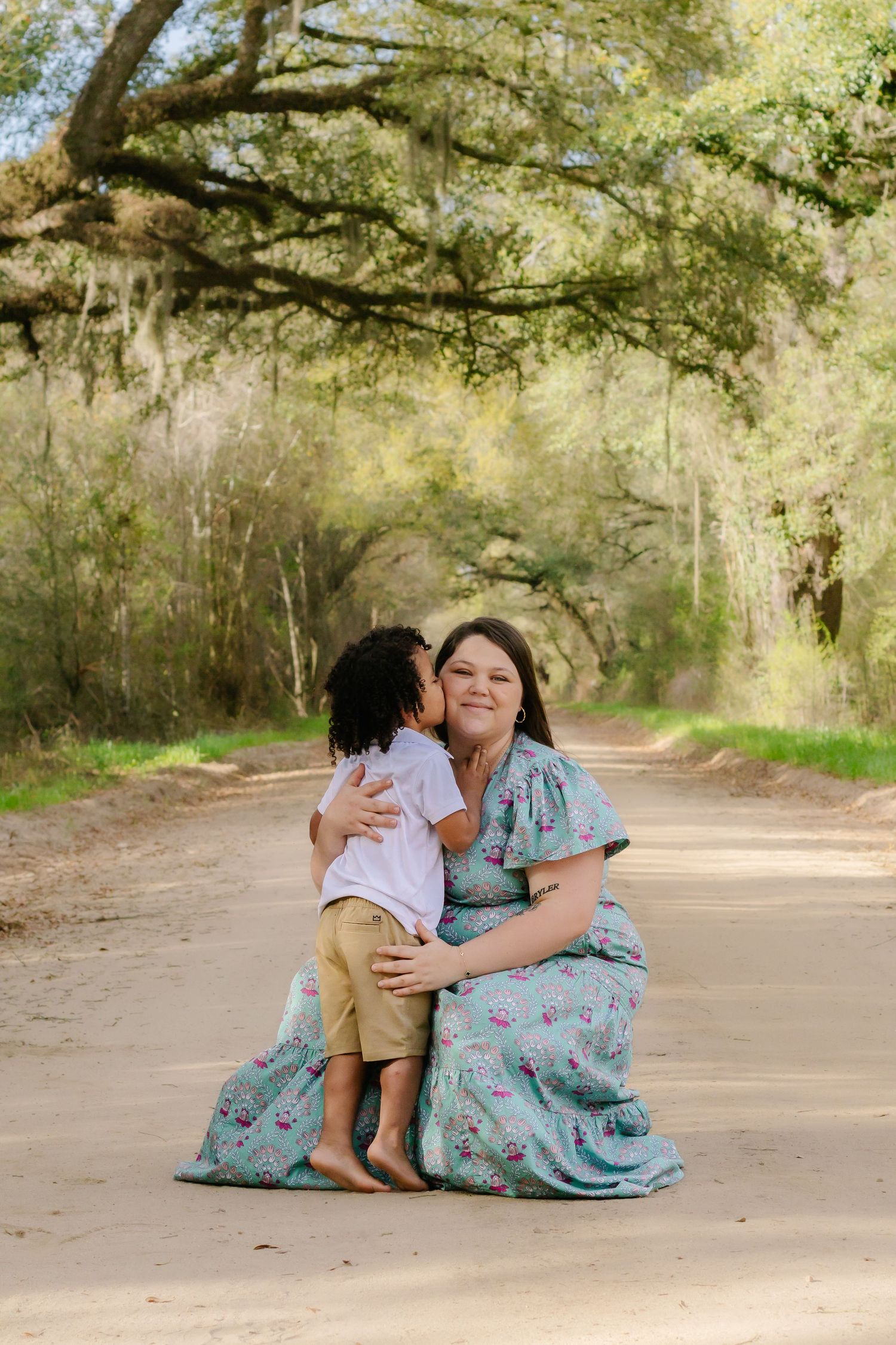 Woman in teal floral dress kneeling on dirt road as young child gives her a kiss on the cheek outdoors.
