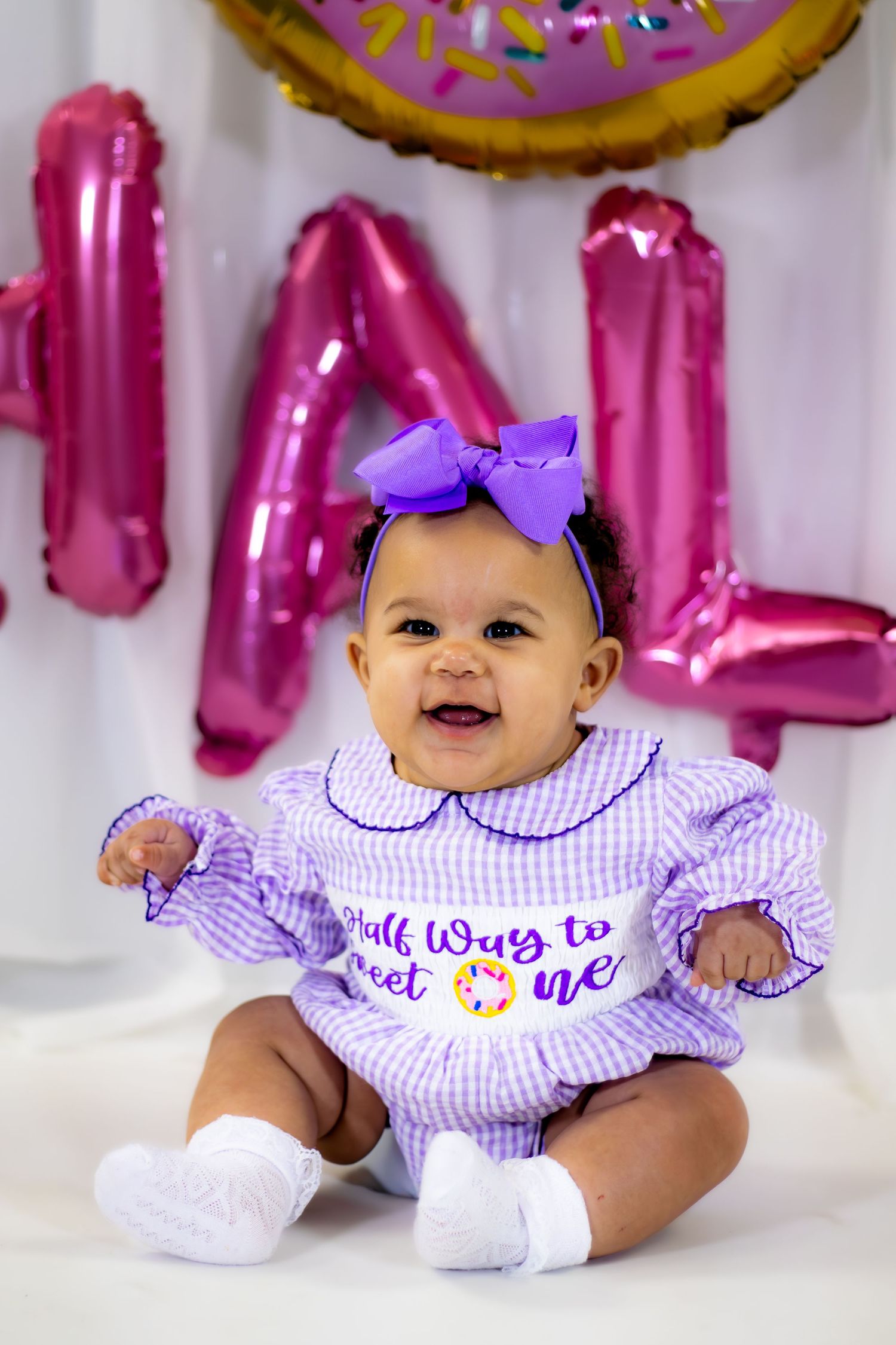 Smiling baby girl in purple gingham outfit with bow headband at donut-themed half birthday celebration.