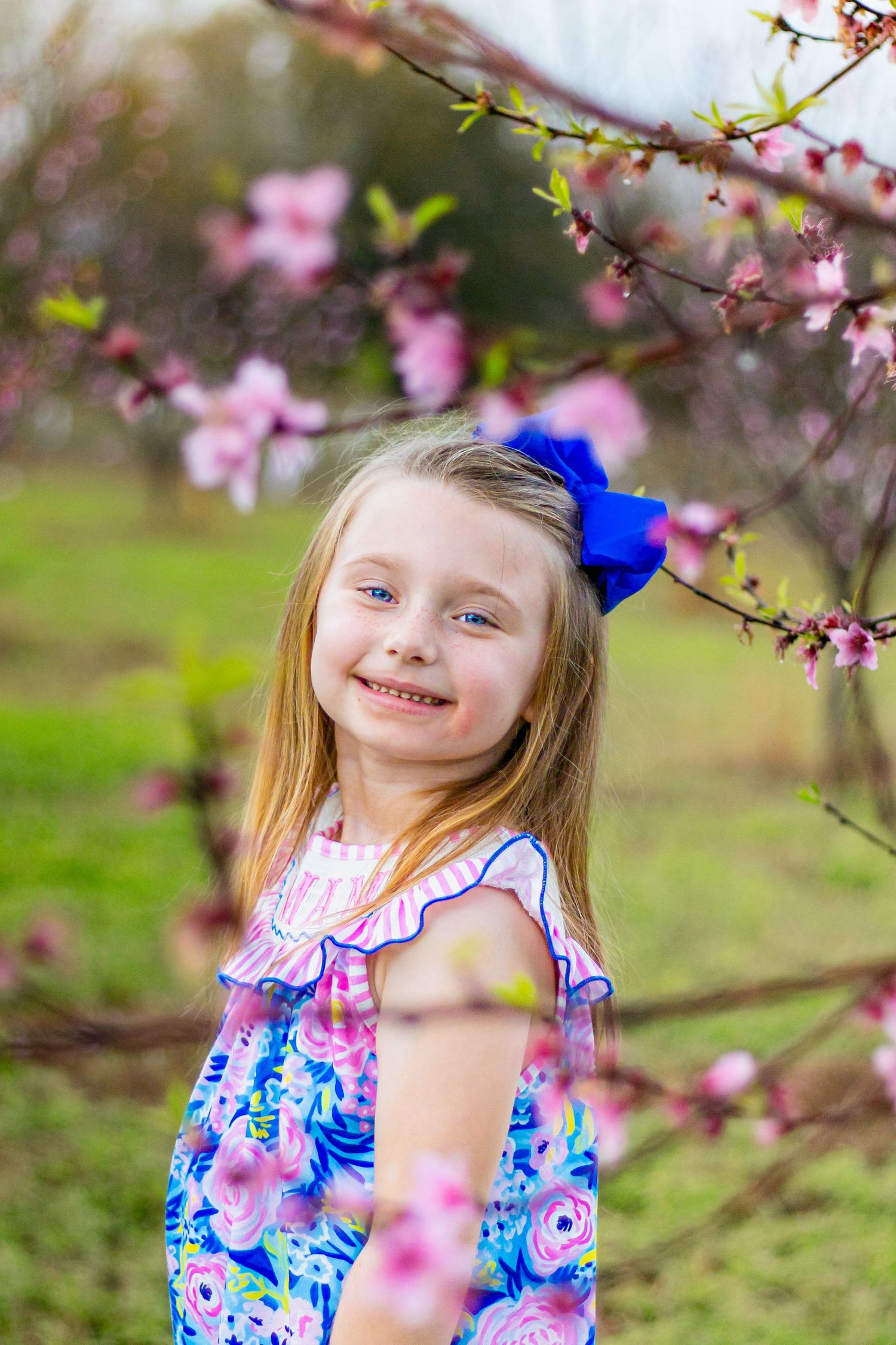Smiling blonde girl with blue bow in hair wearing floral dress stands among pink cherry blossom branches outdoors.