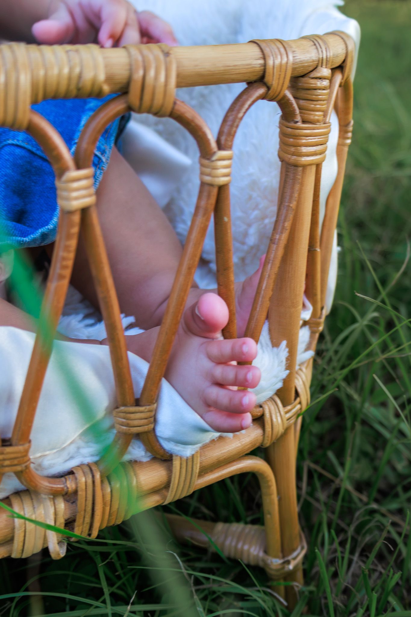 Close-up of a baby's tiny feet peeking through a wicker rattan chair on green grass outdoors.