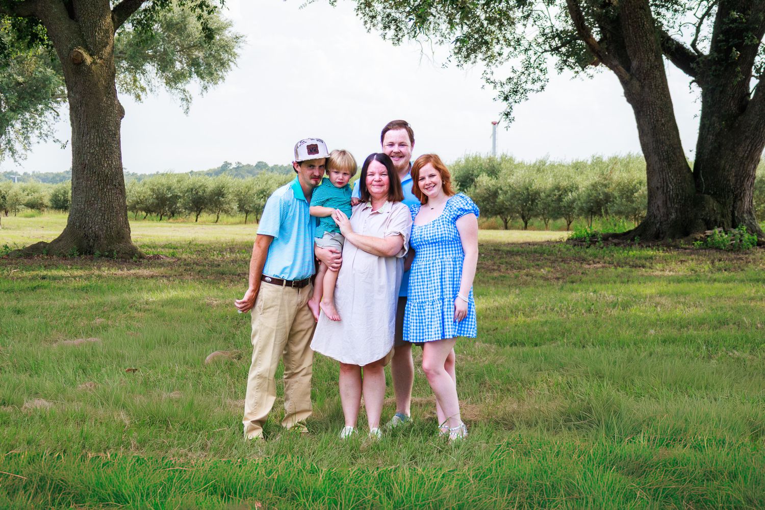 Family of five posing outdoors on green grass under large trees on a cloudy day.