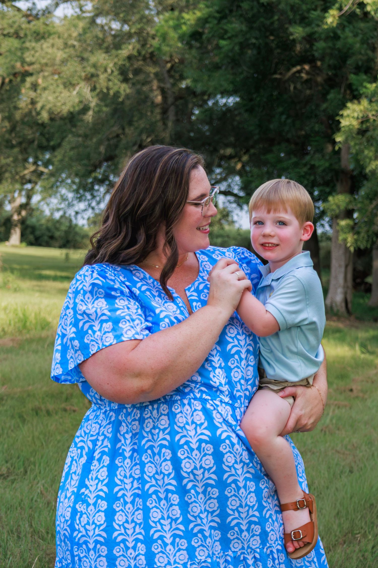 A woman in a blue floral dress holds a smiling toddler boy outdoors in a green park setting.