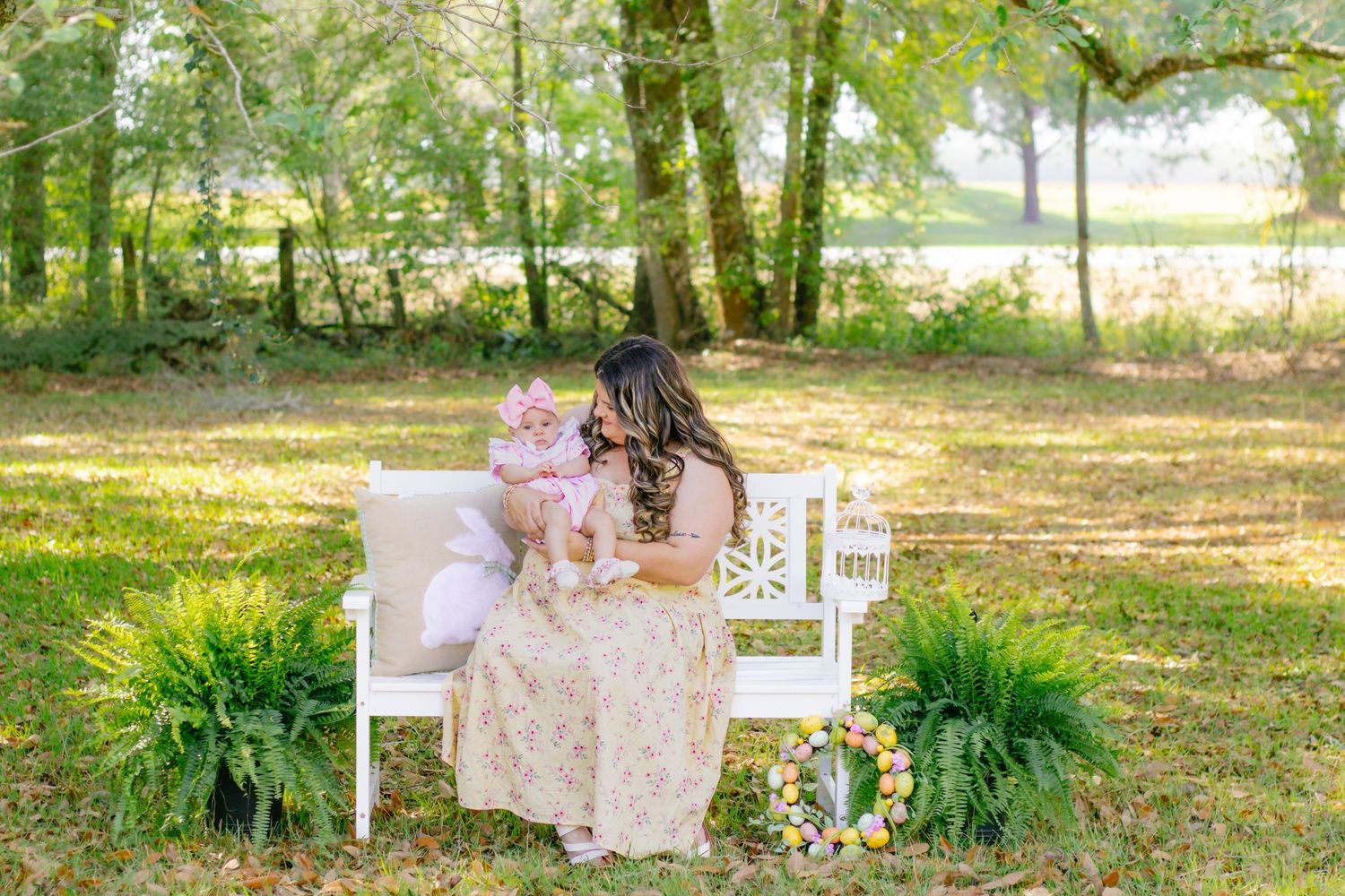 Mother in floral dress sits on white bench holding baby girl in lush green outdoor setting.