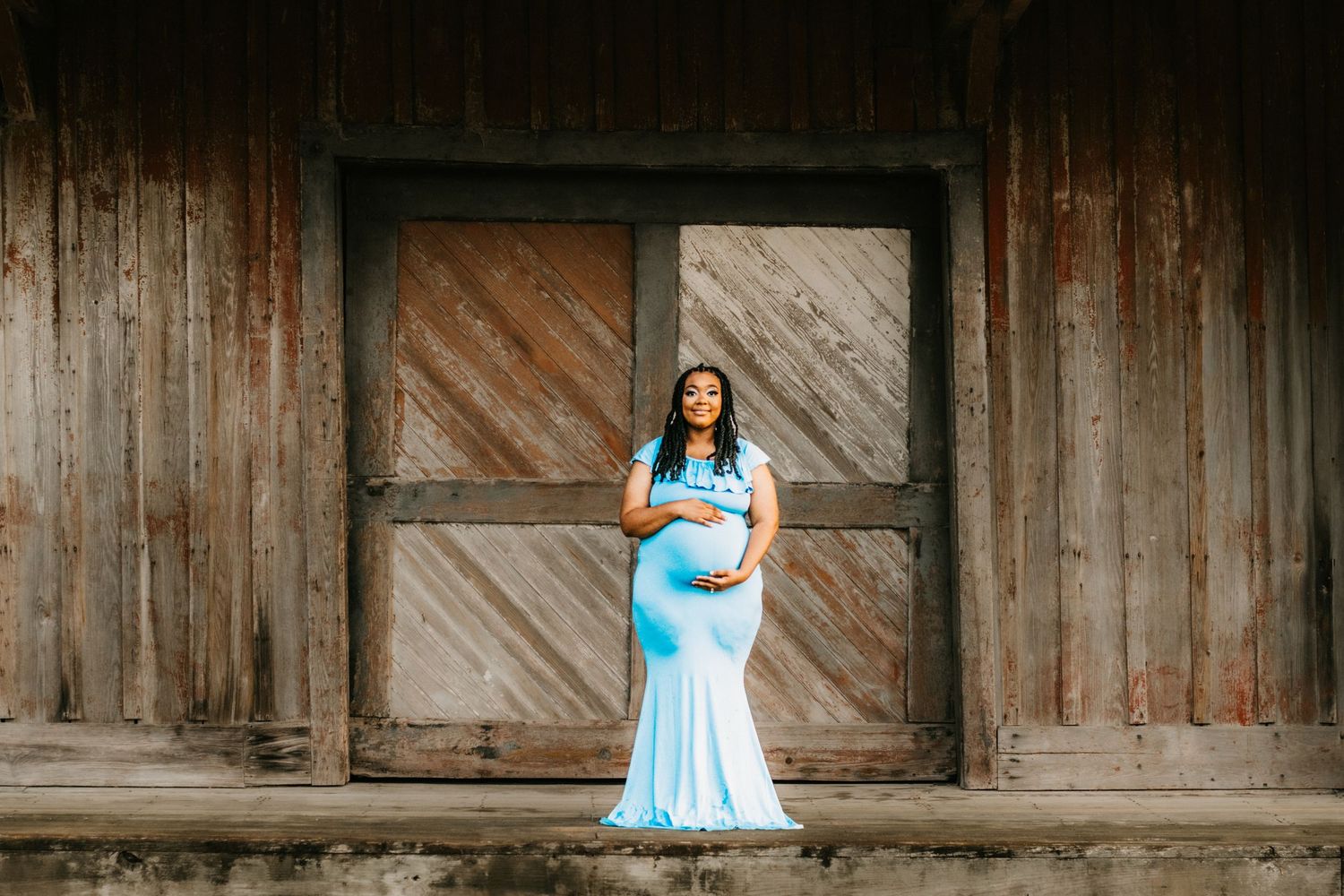 Pregnant woman in elegant light blue mermaid gown poses in front of rustic wooden barn doors.