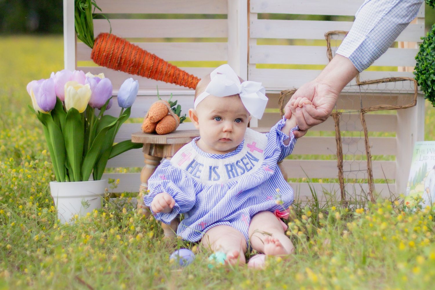 Baby in Easter outfit sits in field with tulips and carrots, holding adult hand.