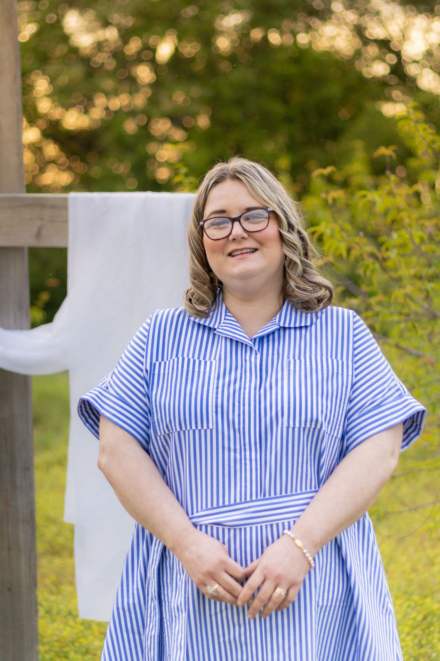 Cheerful woman with glasses wearing blue striped dress smiling outdoors near a wooden cross with white draping.