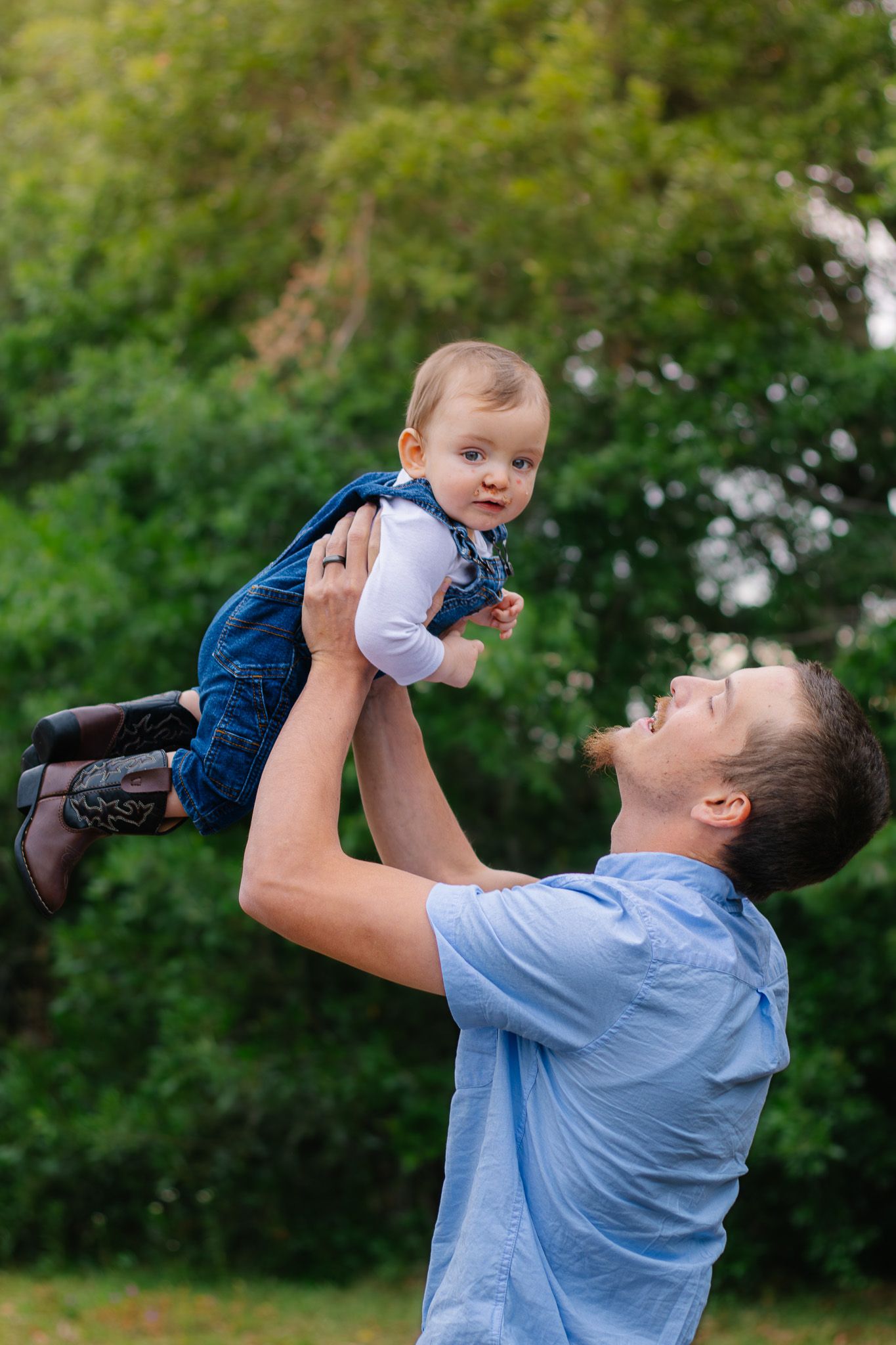 Loving father holding baby in denim overalls up in the air while smiling in outdoor setting.