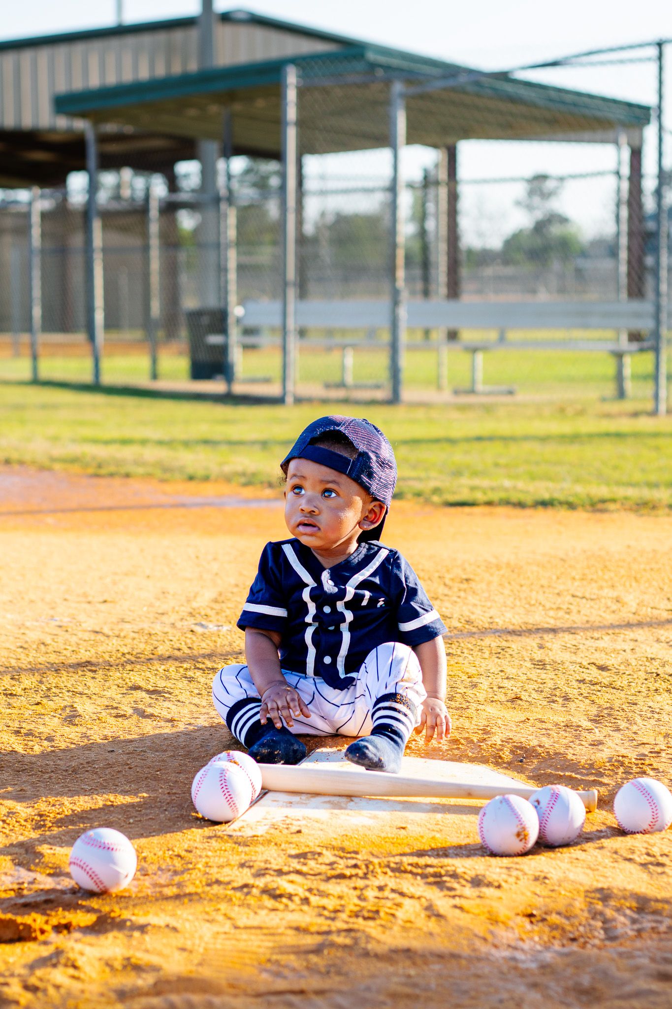 Toddler in baseball uniform sitting on dirt field with bats and balls.
