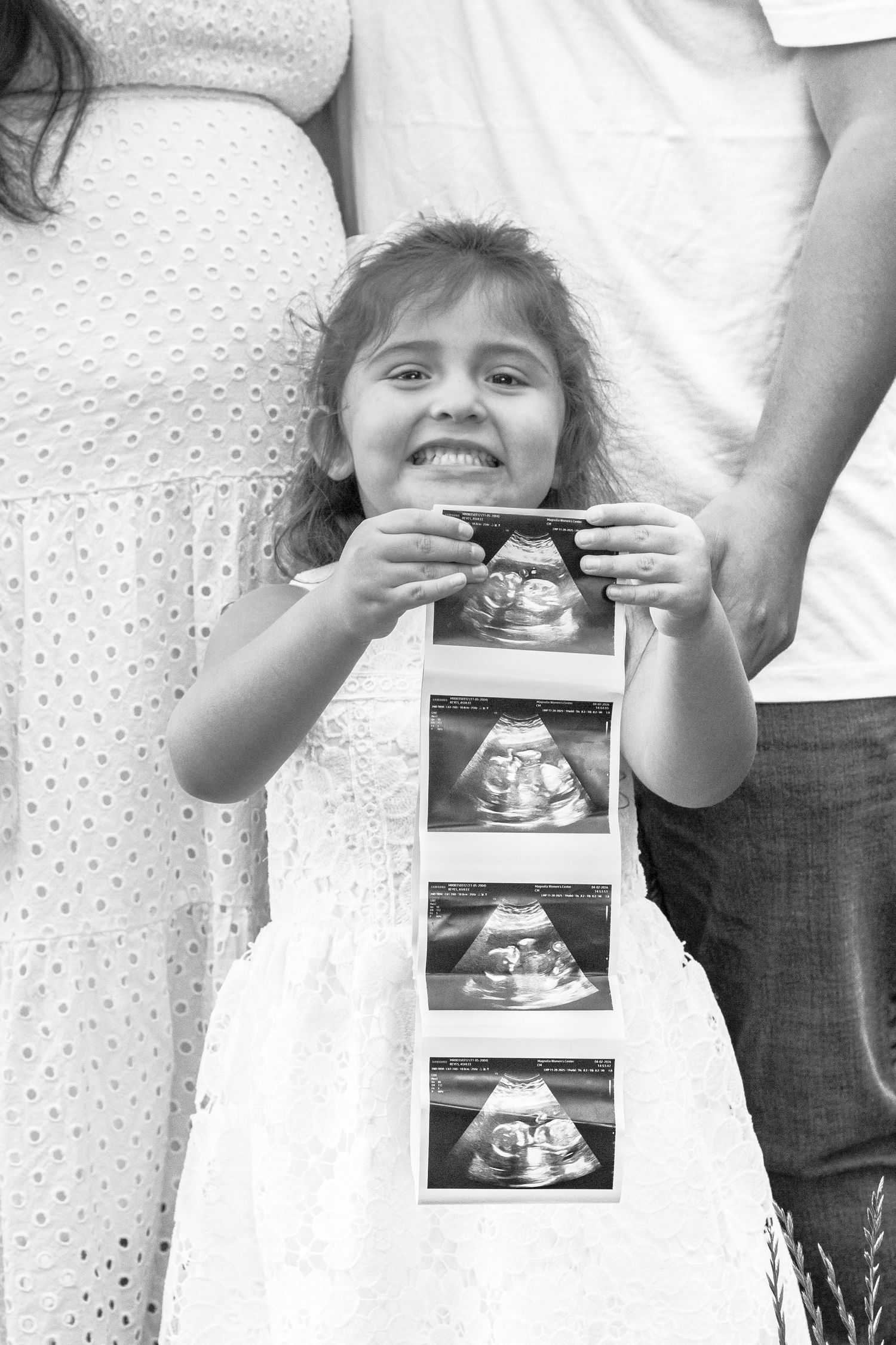 A smiling girl in white dress holds ultrasound photos between her parents during a pregnancy announcement photoshoot.