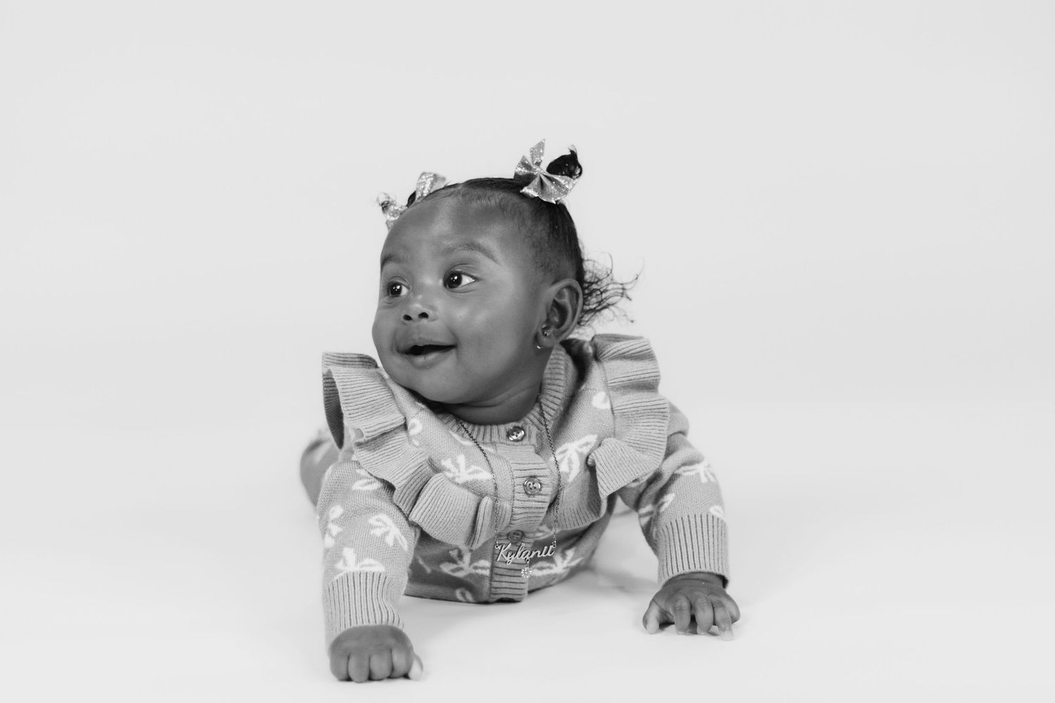 Black and white photo of a smiling baby girl lying on her stomach, wearing a ruffled outfit with hair bows.