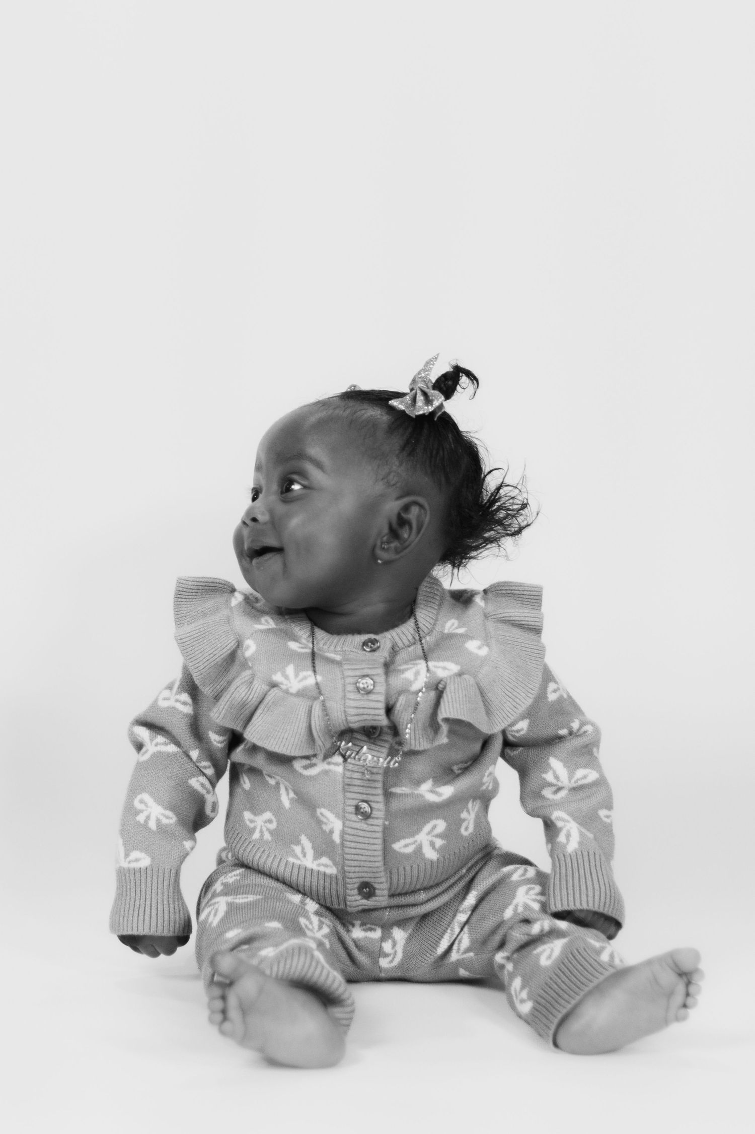 Black and white photo of an adorable baby girl sitting up, wearing a ruffled patterned outfit with a small hair bow.