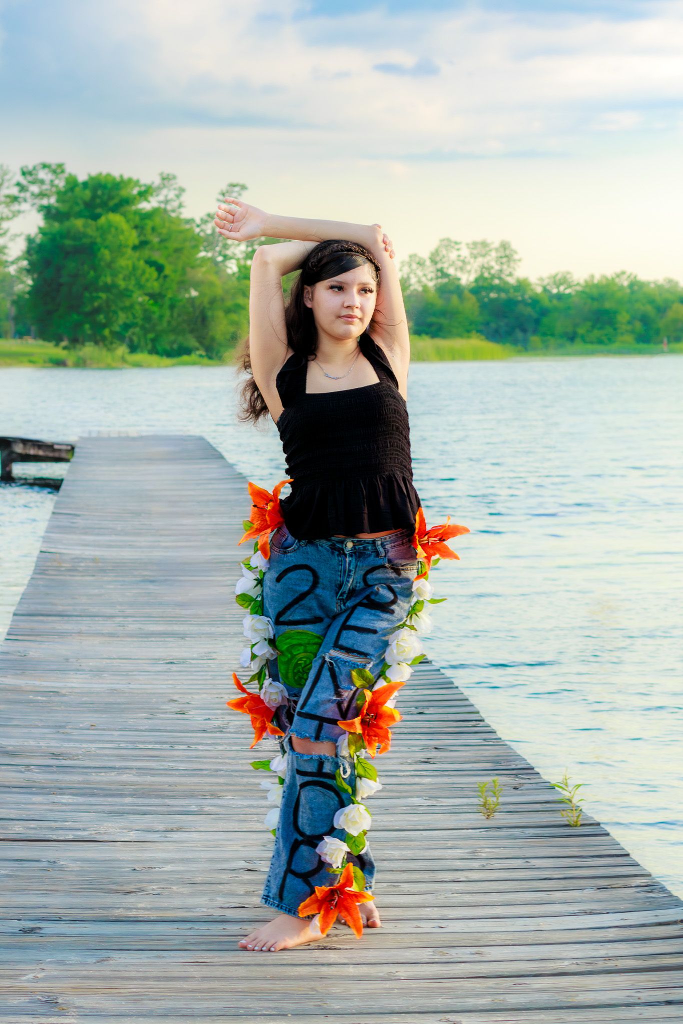 Young woman in black top and floral jeans poses on wooden dock by lake at sunset.