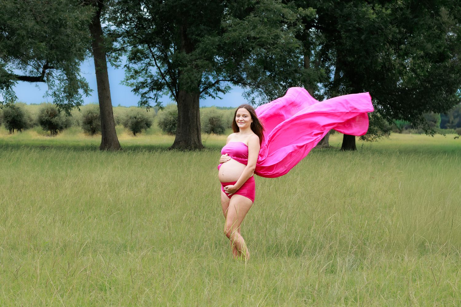 Pregnant woman in pink bikini and flowing pink fabric poses in lush green field with trees in background.