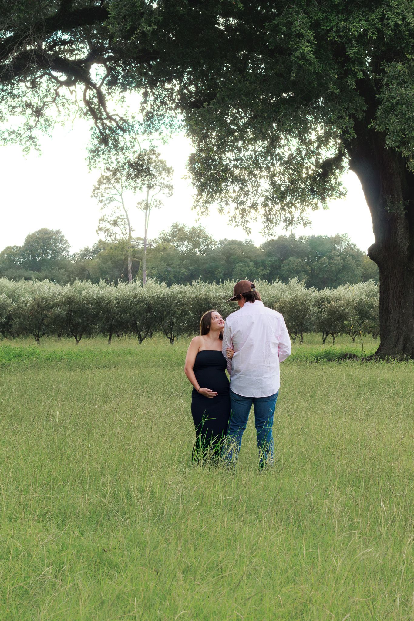 Pregnant couple standing in lush green field under large oak tree during outdoor maternity photo session.