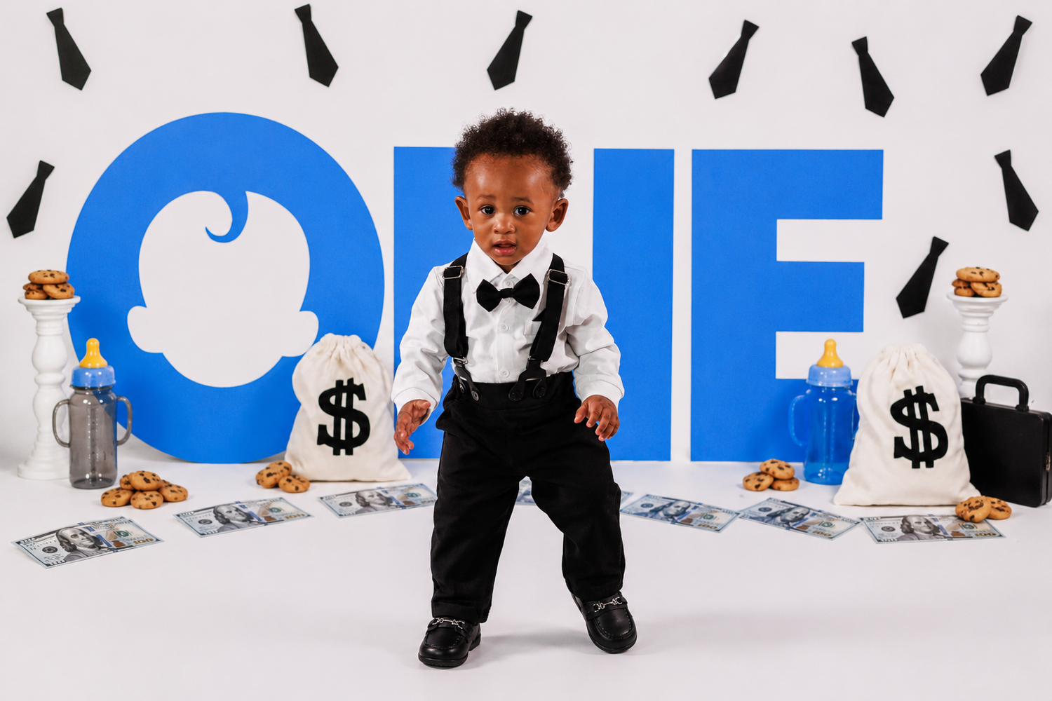 Toddler boy in suspenders and bow tie poses before a blue ONE backdrop with dollar signs for a first birthday photo.