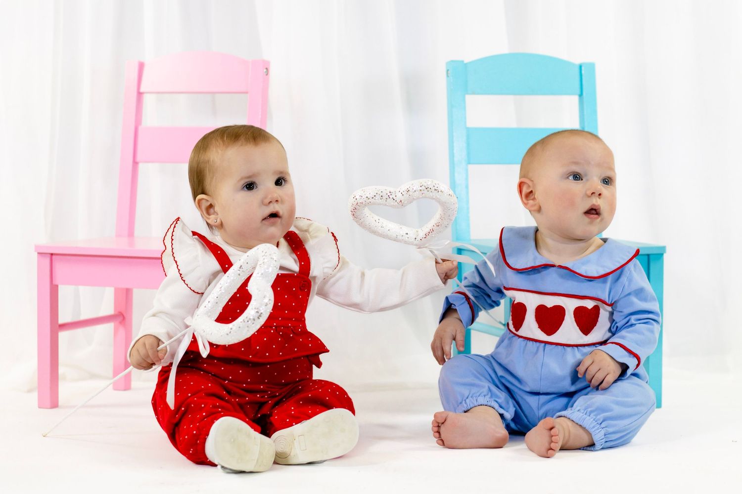 Two babies in Valentine's Day outfits sit together holding a heart wand between pink and blue chairs.