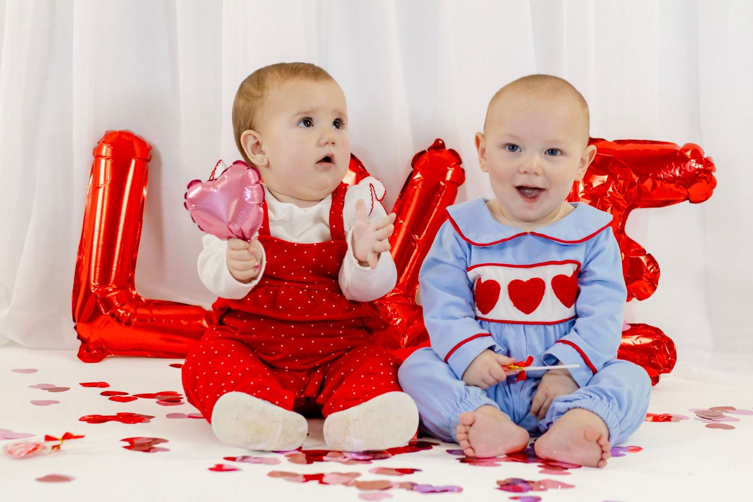 Two babies in Valentine outfits sit before a red LOVE balloon with heart confetti scattered around.