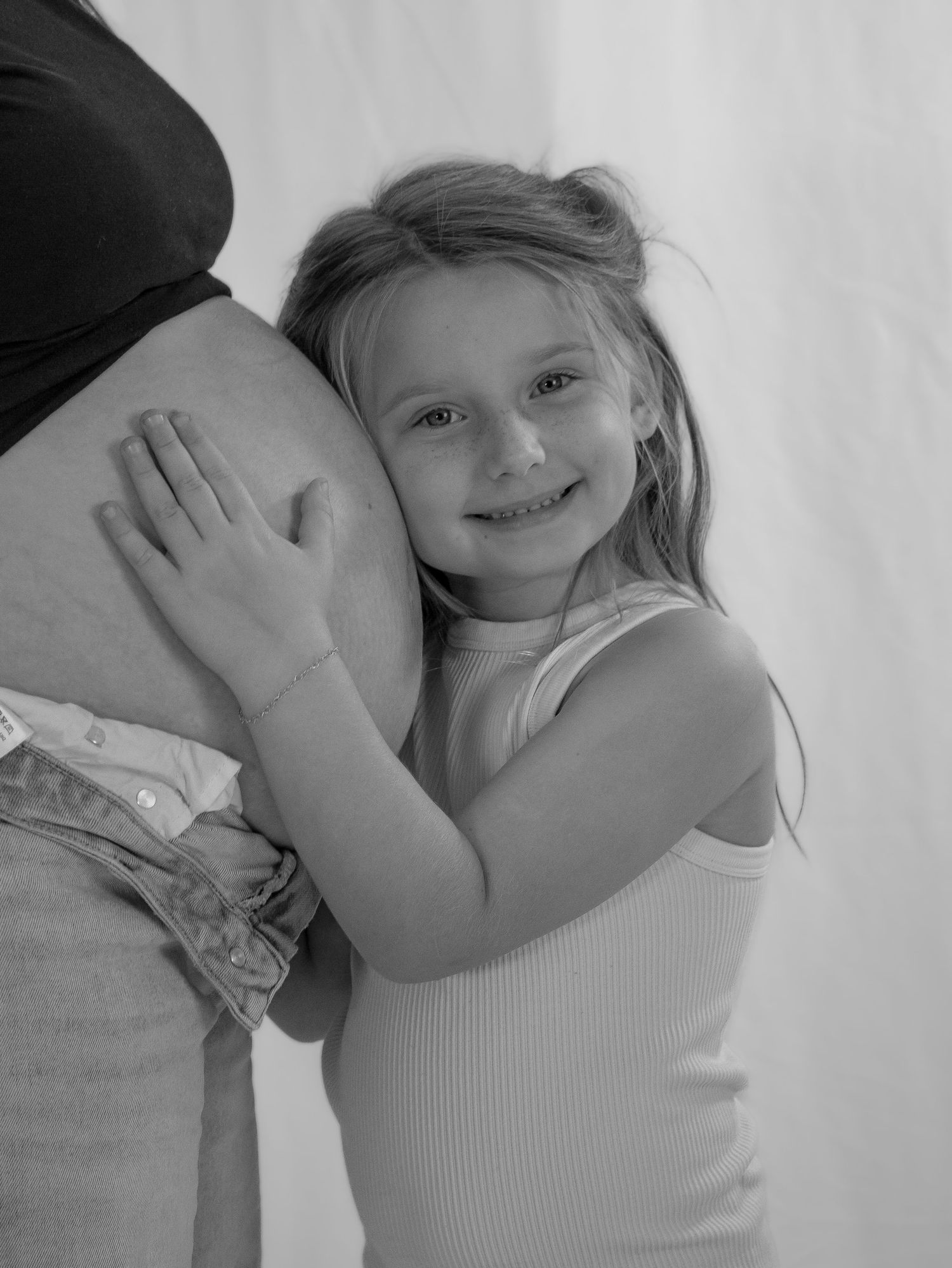 Black and white photo of smiling young girl hugging a pregnant womans baby bump tenderly.