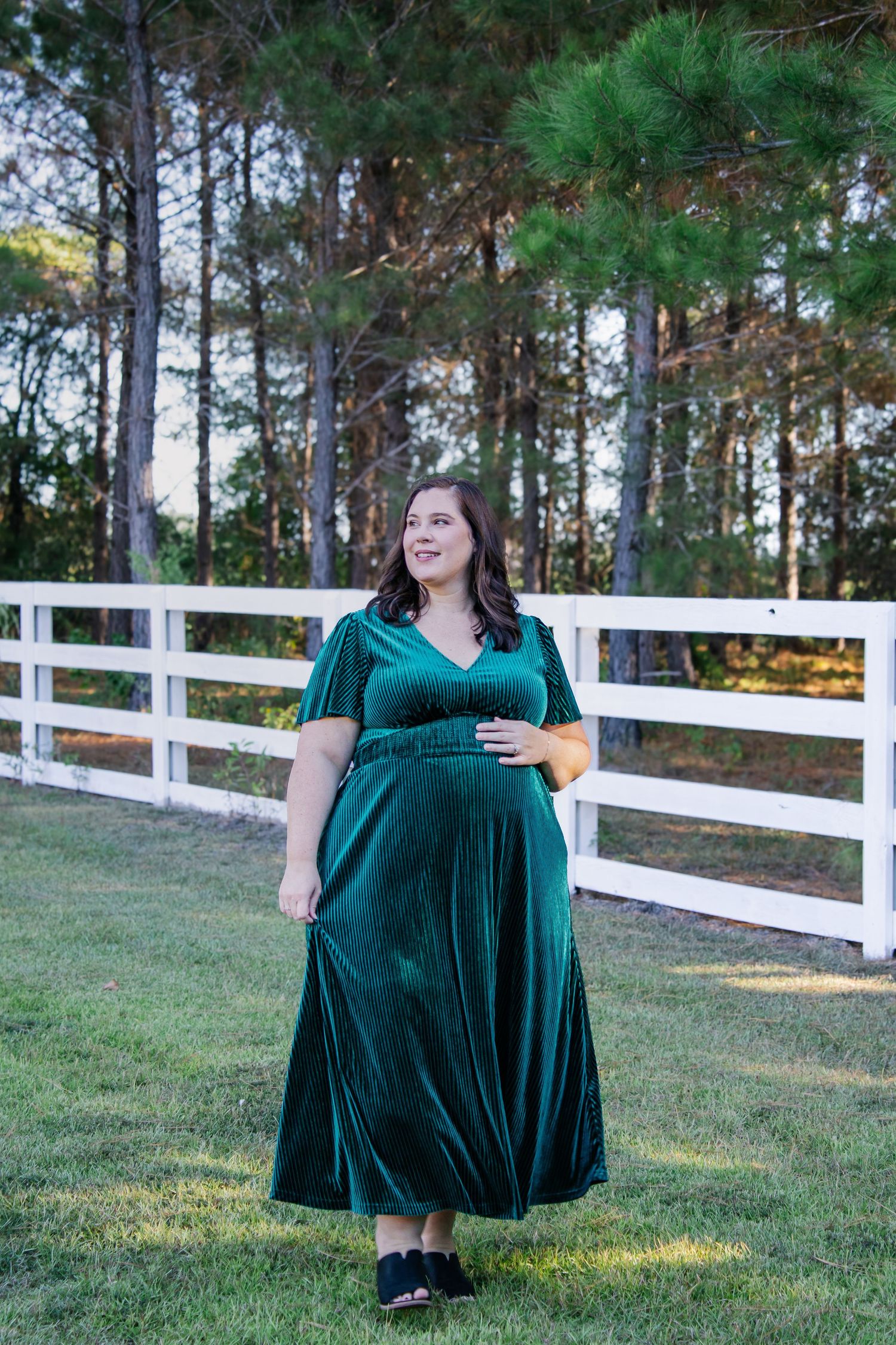 Pregnant woman in emerald velvet dress standing by white fence near pine trees.
