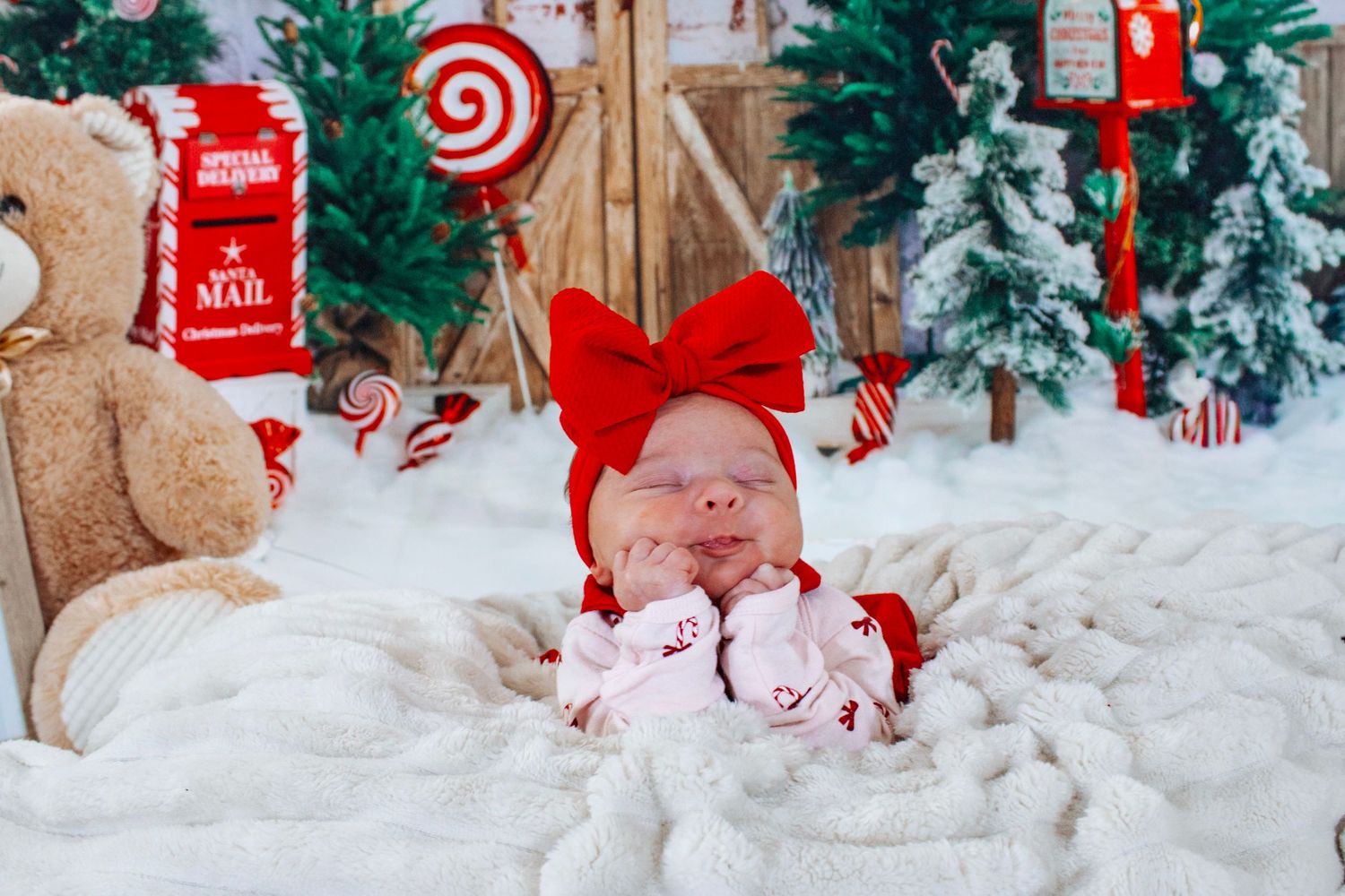 A newborn baby wearing a red bow headband sleeps peacefully in a festive Christmas-themed setting.