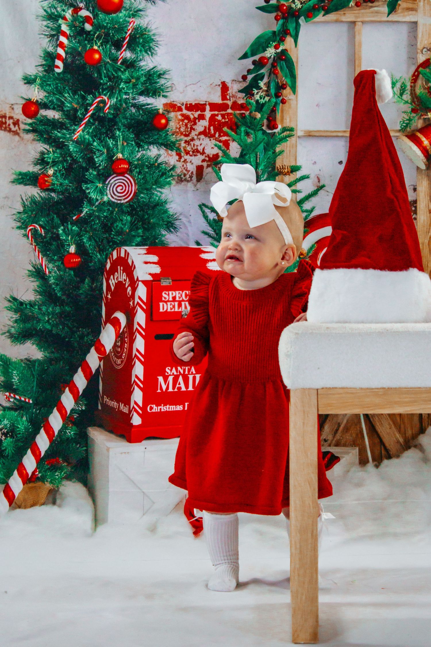 A baby girl in a red dress reaches toward a Santa hat beside a decorated Christmas tree indoors.