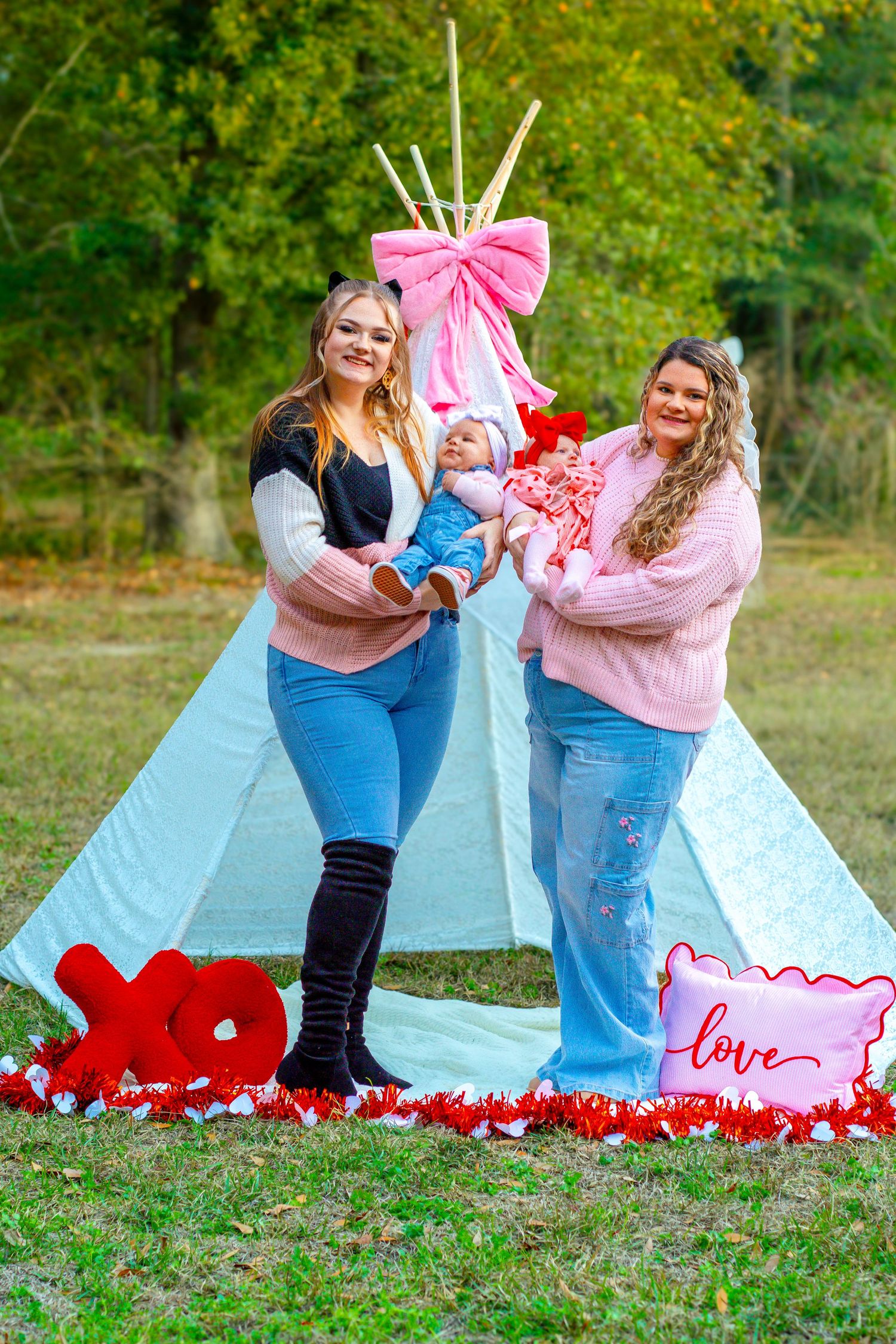 Two women holding babies stand before white teepee decorated with pink bows and Valentine XO and love signs.