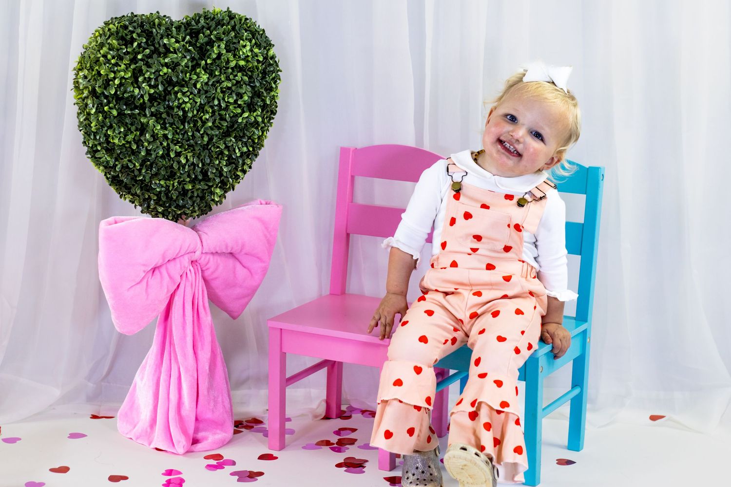 Happy toddler in heart-print overalls sits on a blue chair beside a heart-shaped topiary with pink bow.