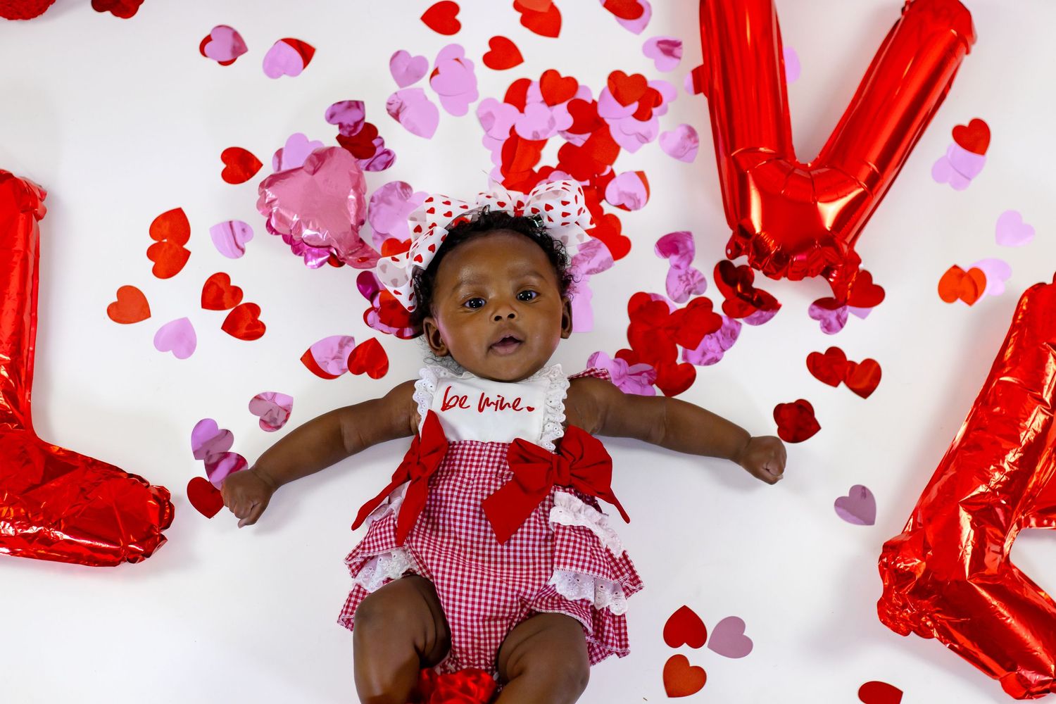 Newborn baby in red gingham dress laying among heart confetti with LOVE balloons for Valentine's Day.