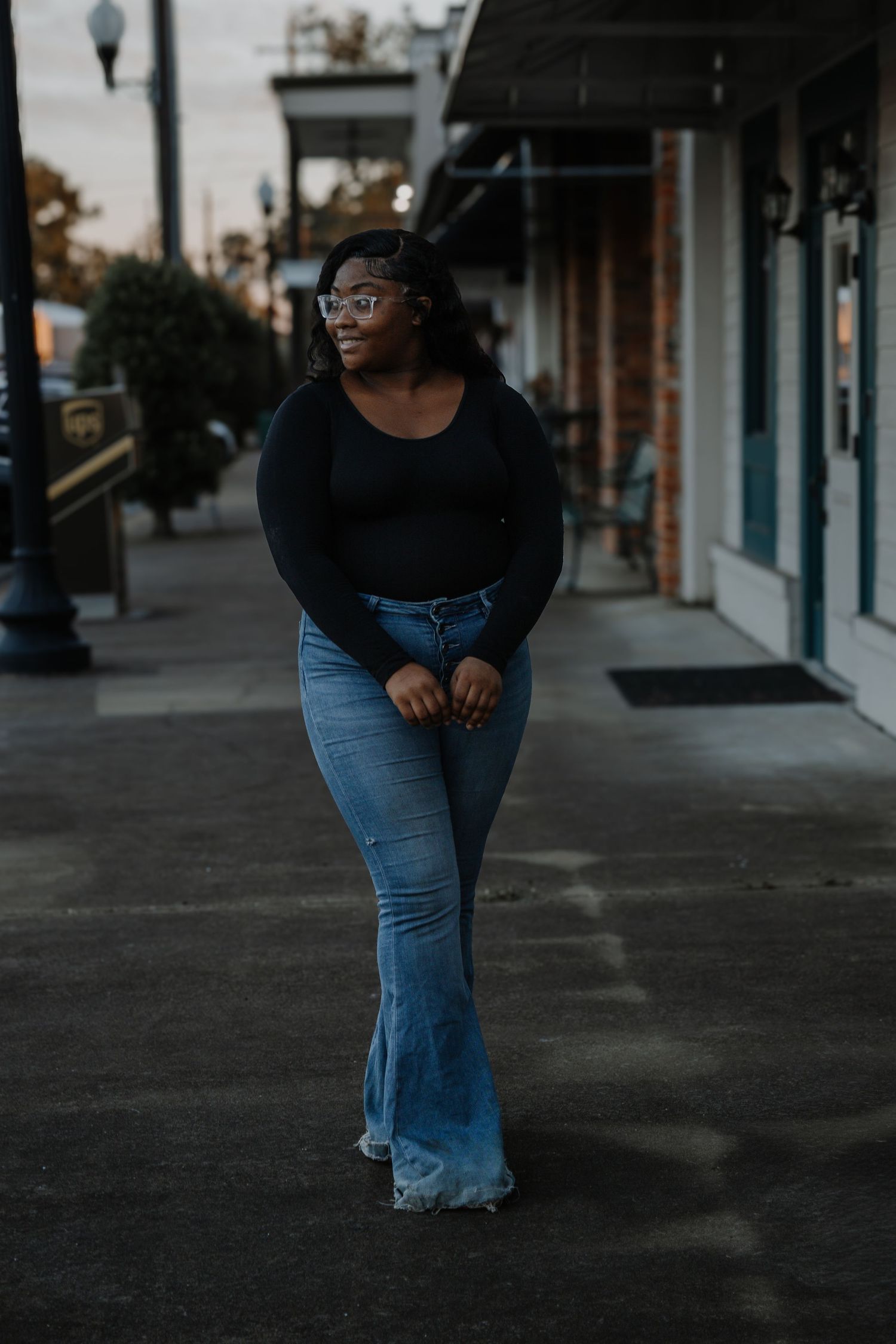 Young Black woman in black bodysuit and flared jeans walks along a small town sidewalk.