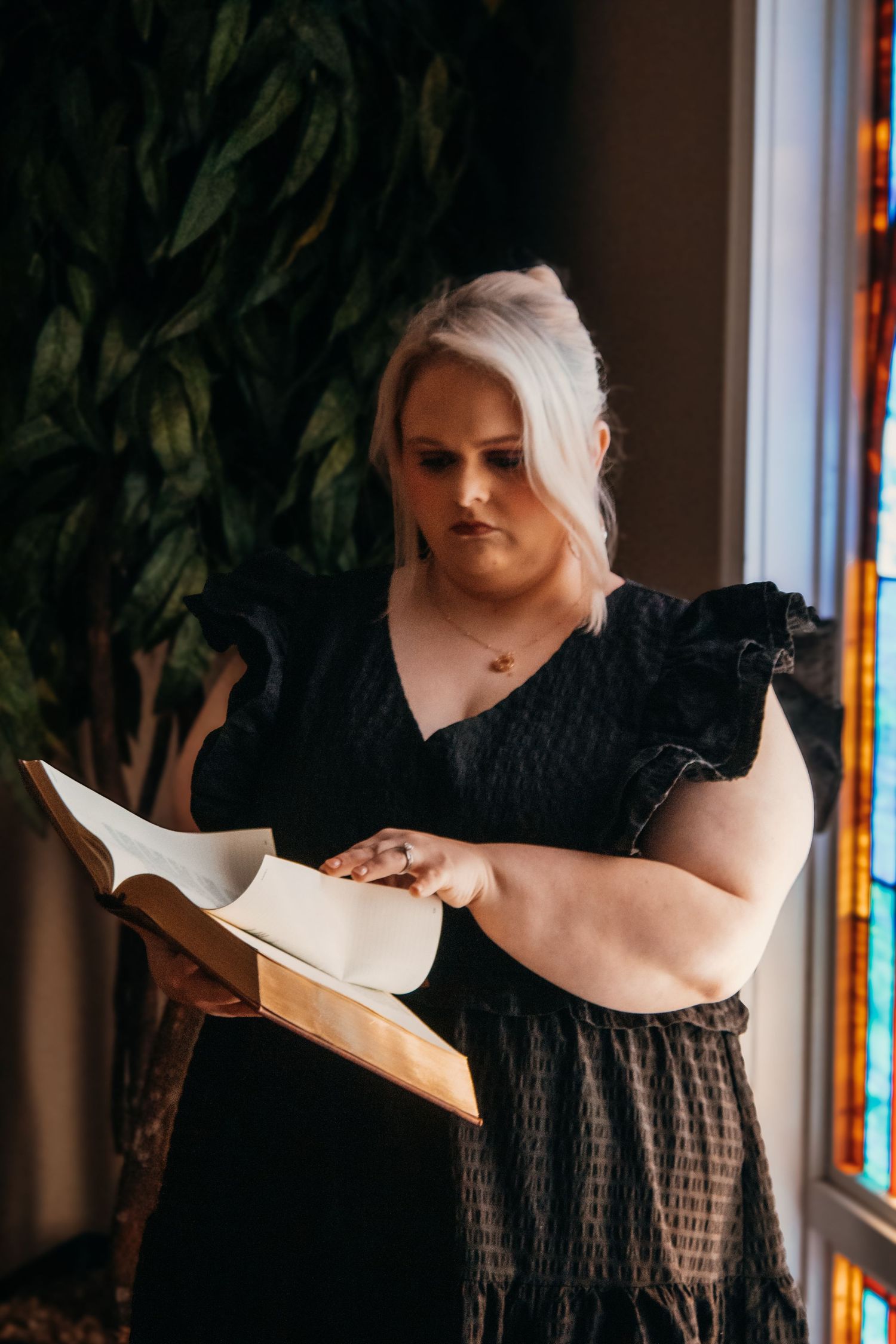 Blonde woman in black ruffled dress reading an open book near a stained glass window in a church.