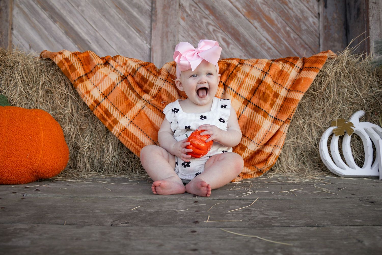 Laughing baby girl in floral romper with pink bow sitting on plaid blanket with fall pumpkins.