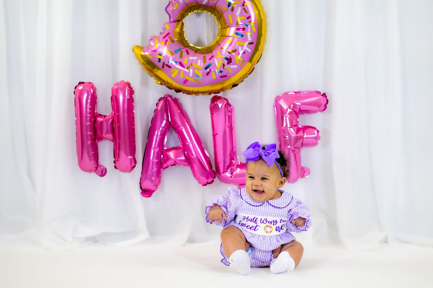 Baby girl in purple romper celebrates half birthday with pink HALF balloons and donut balloon backdrop.