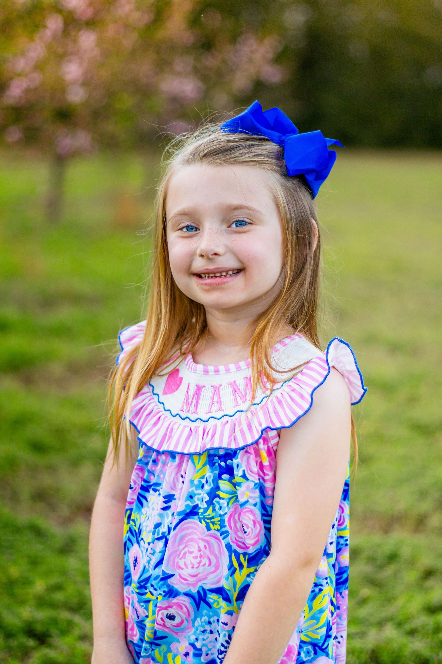 Cheerful blonde girl in colorful floral dress with blue hair bow smiles in a green field with blooming trees.