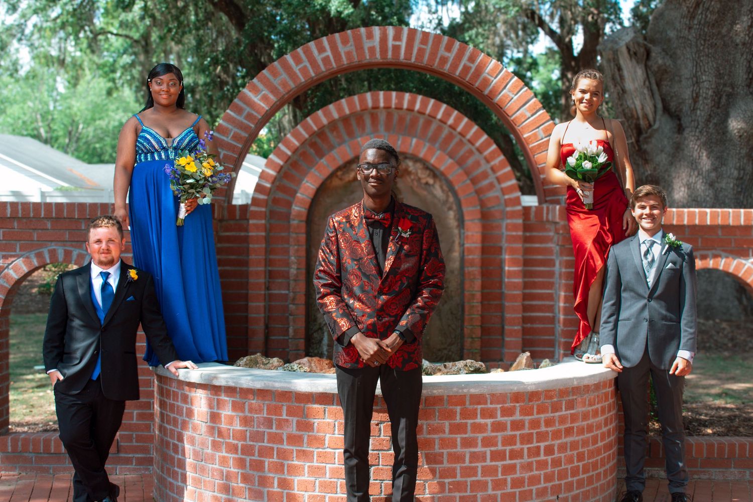 Four prom students pose around a decorative brick well, dressed in elegant formal attire for their special night.