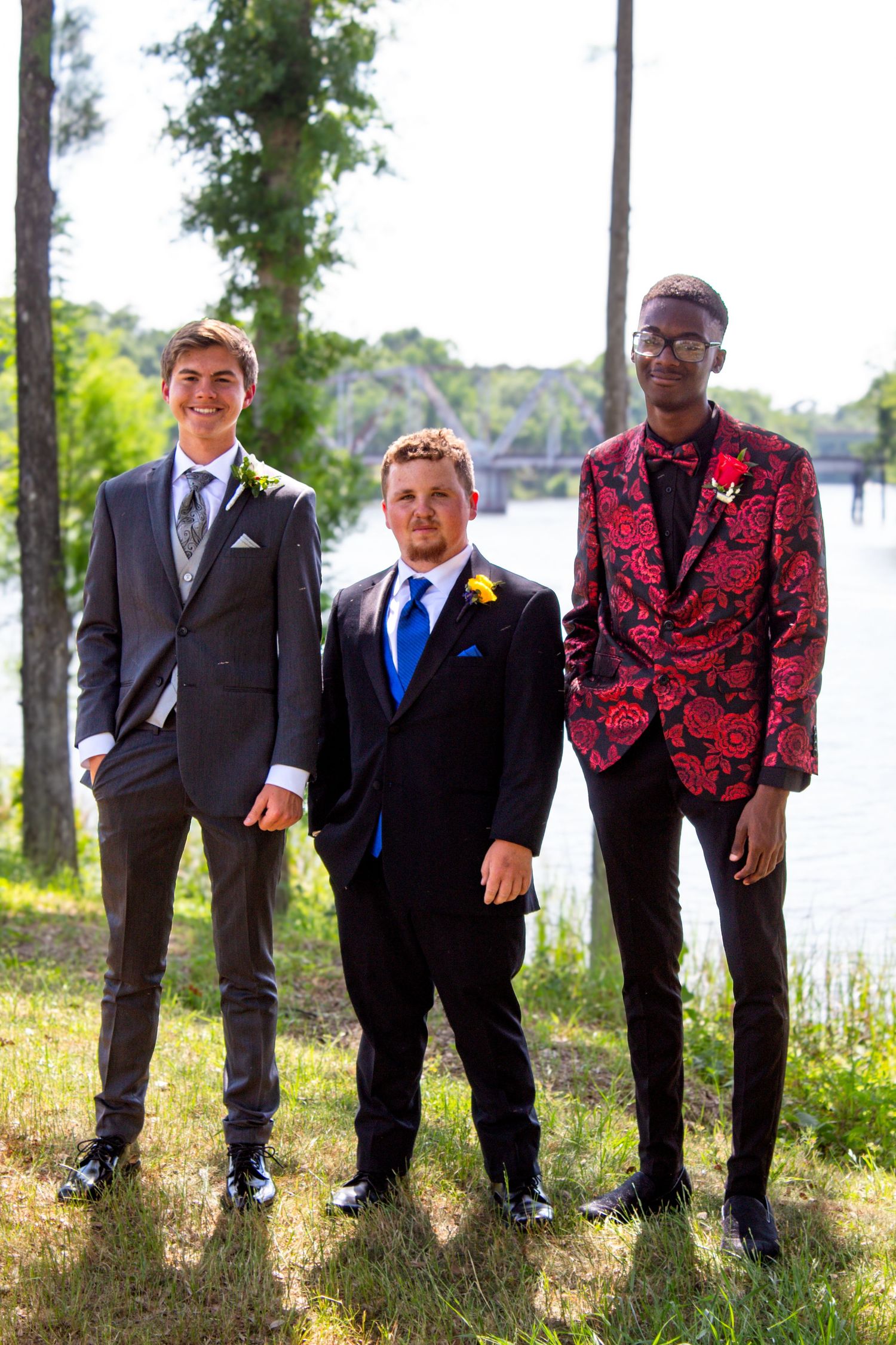 Three young men in formal prom attire pose outdoors near a tree-lined river.