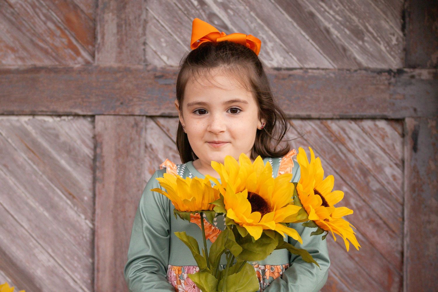 Adorable young girl with orange bow holds bright yellow sunflowers in front of rustic wooden barn door.