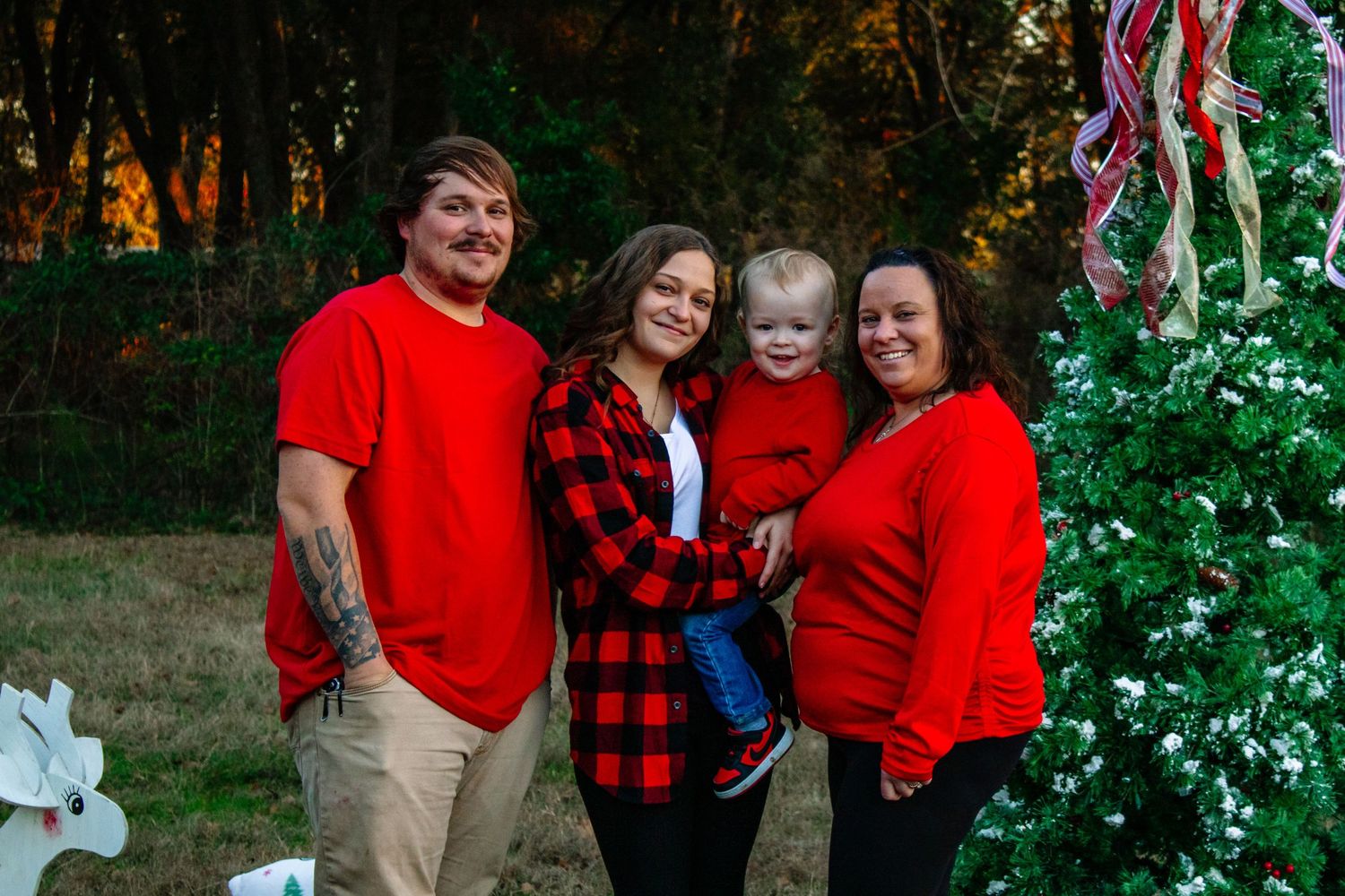 Family of four wearing red outfits smiling together beside a decorated outdoor Christmas tree in the evening light.